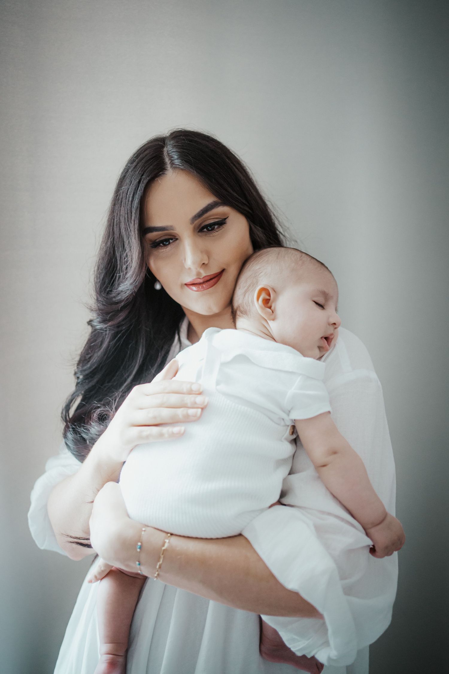 Lächelnde, dunkelhaarige Mutter im weißen Kleid hält ihr schlafendes Baby sanft an ihrer Schulter im natürlichen Licht.