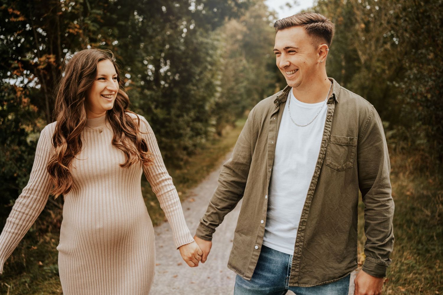 Smiling couple holding hands and walking along a path in the woods, the woman in a fitted beige dress.