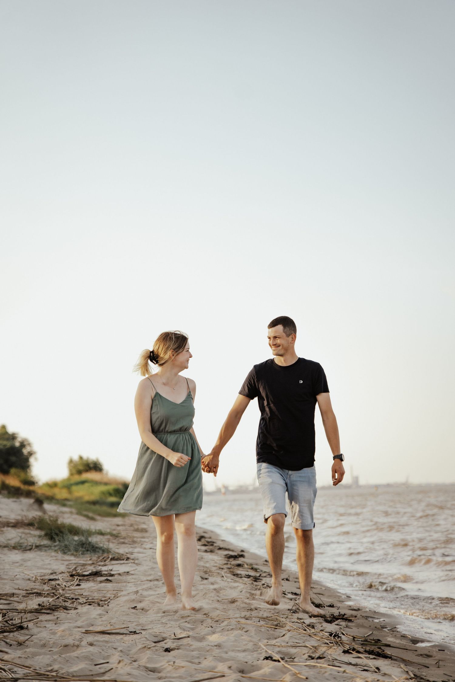 Barefoot couple holding hands and smiling as they stroll along a sandy beach shoreline at sunset.