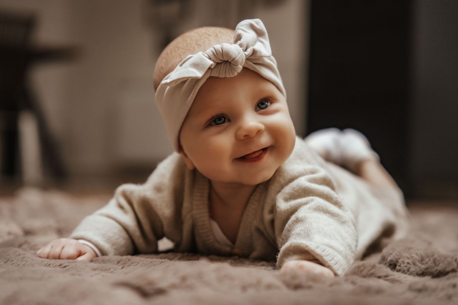 Smiling baby girl in a beige knotted headband lying on her tummy on a soft textured rug at home.
