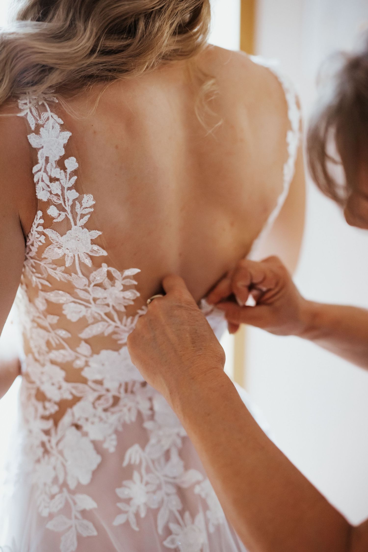 Detailaufnahme von Händen, die die Knöpfe am Rücken eines Brautkleides mit floraler Spitze schließen.