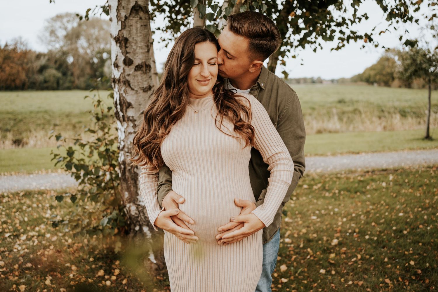 Man kissing his pregnant partner's cheek as they cradle her baby bump beside a birch tree.