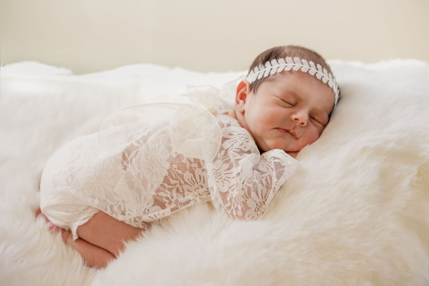 Sleeping newborn baby in a white lace romper and sparkly headband resting peacefully on a fluffy white sheepskin.