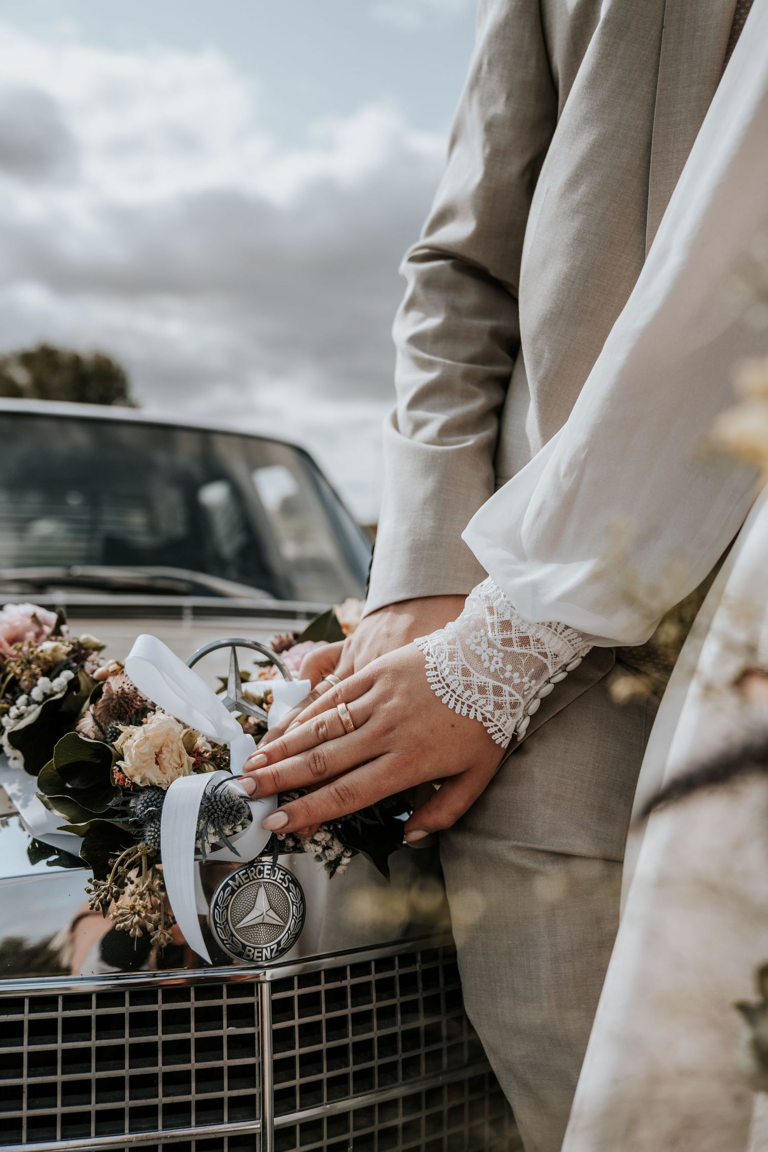 Detailaufnahme der Hände des Brautpaares vor einem Oldtimer mit Trockenblumen. Die Eheringe und der Spitzenärmel der Braut sind sichtbar.
