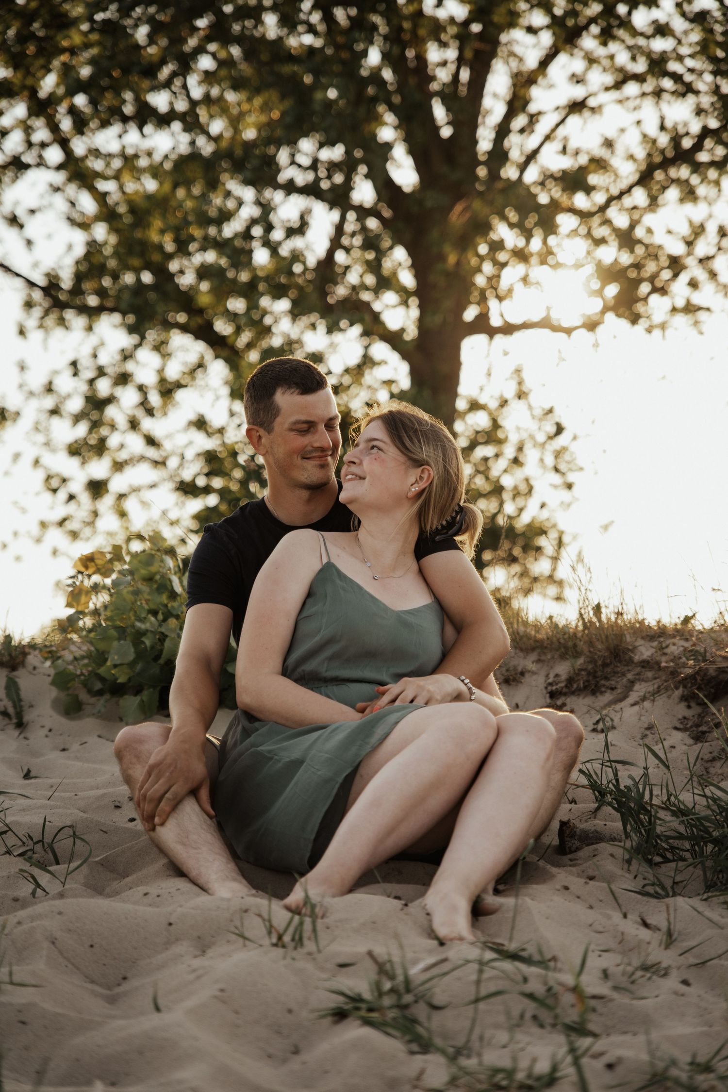 Affectionate couple cuddling on a sandy dune beneath a large tree in warm evening light.