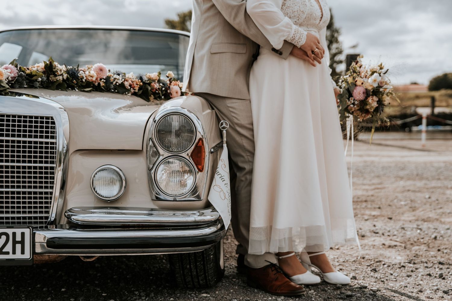 Brautpaar umarmt sich neben einem klassischen Mercedes, der bei einer rustikalen Hochzeit mit einer Blumengirlande geschmückt ist. Nordenham