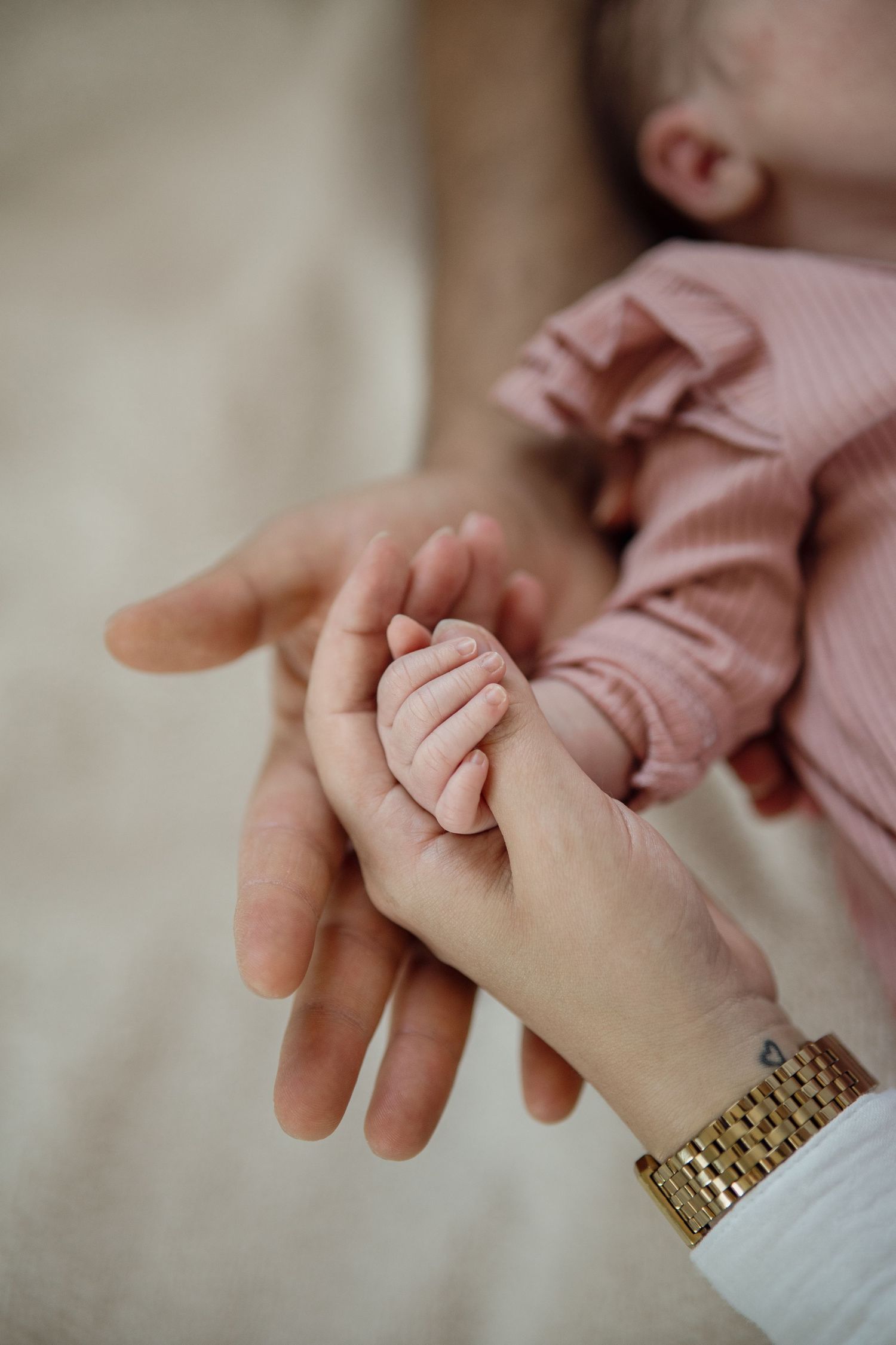 Hände der Eltern halten die winzige Hand eines Neugeborenes. Das Baby trägt ein rosafarbenes Rüschenkleid.
