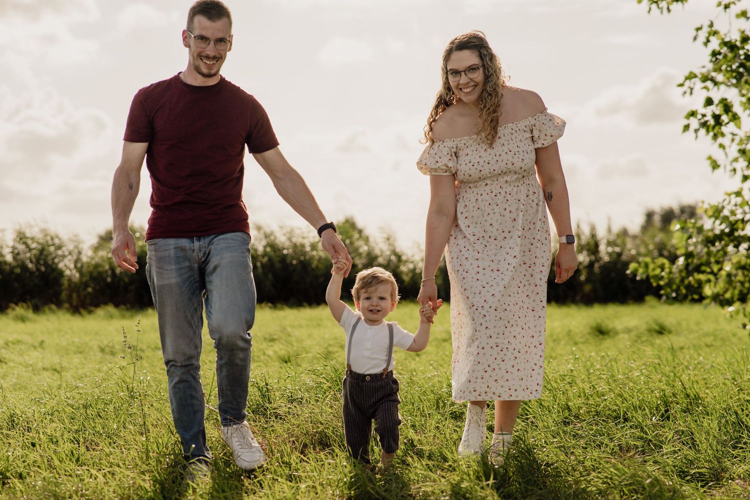 Familie mit Mutter, Vater und kleinem Sohn hält Händchen und spaziert gemeinsam über eine sonnige grüne Wiese.