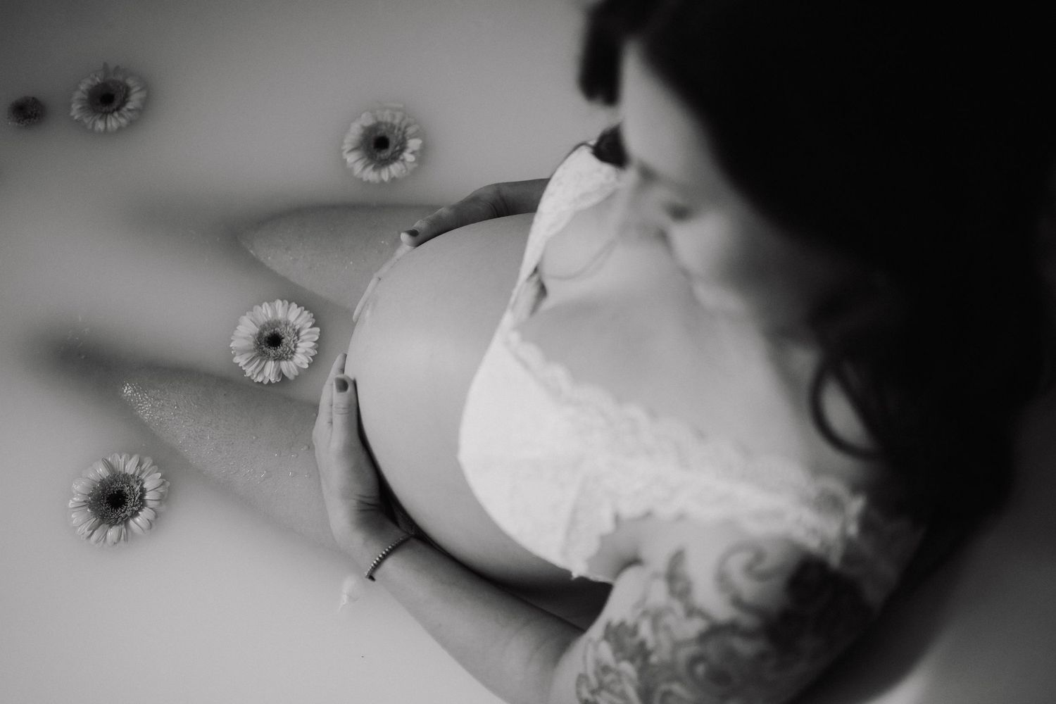 Schwarz-Weiß-Aufnahme einer schwangeren Frau in einer Milchbadewanne, die ihren Bauch hält, umgeben von Gerbera-Blüten.