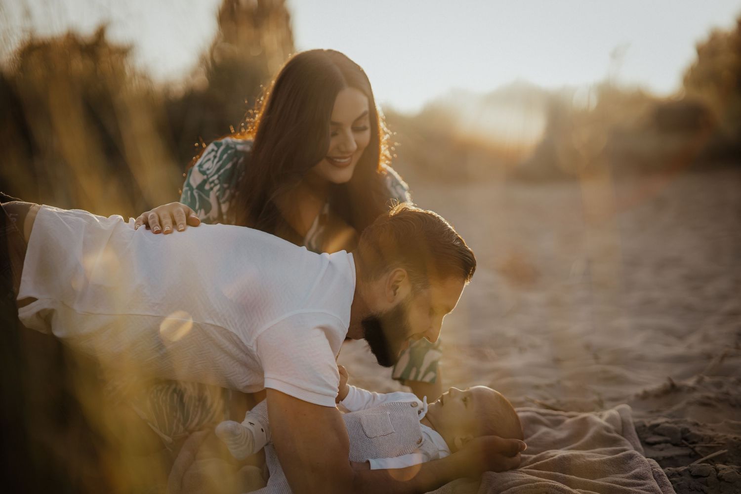 Mutter und Vater kuscheln mit ihrem Neugeborenen im Sand und genießen das warme, goldene Abendlicht.