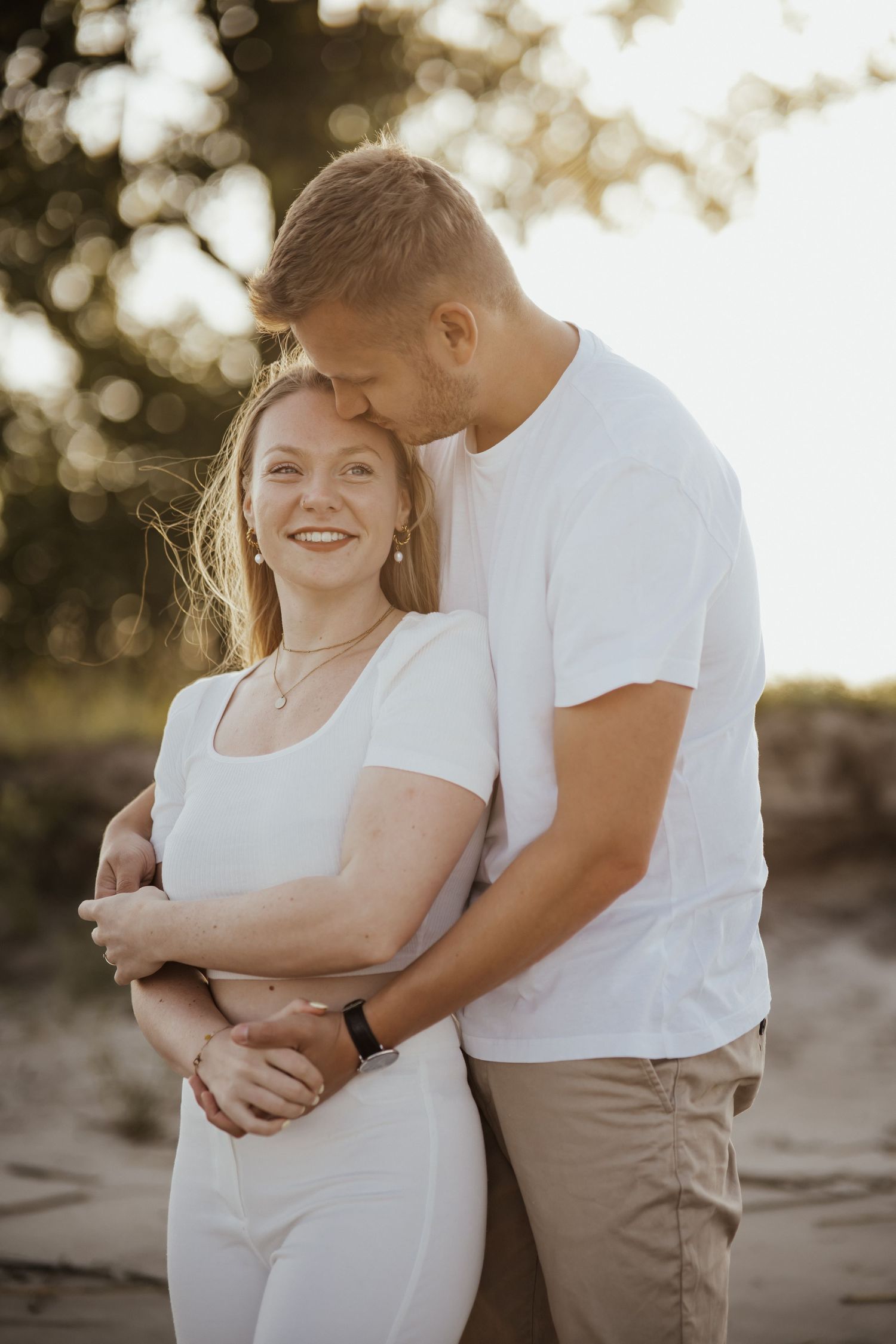 A man in a white t-shirt kisses the forehead of a smiling woman during a golden hour couple photoshoot outdoors.
