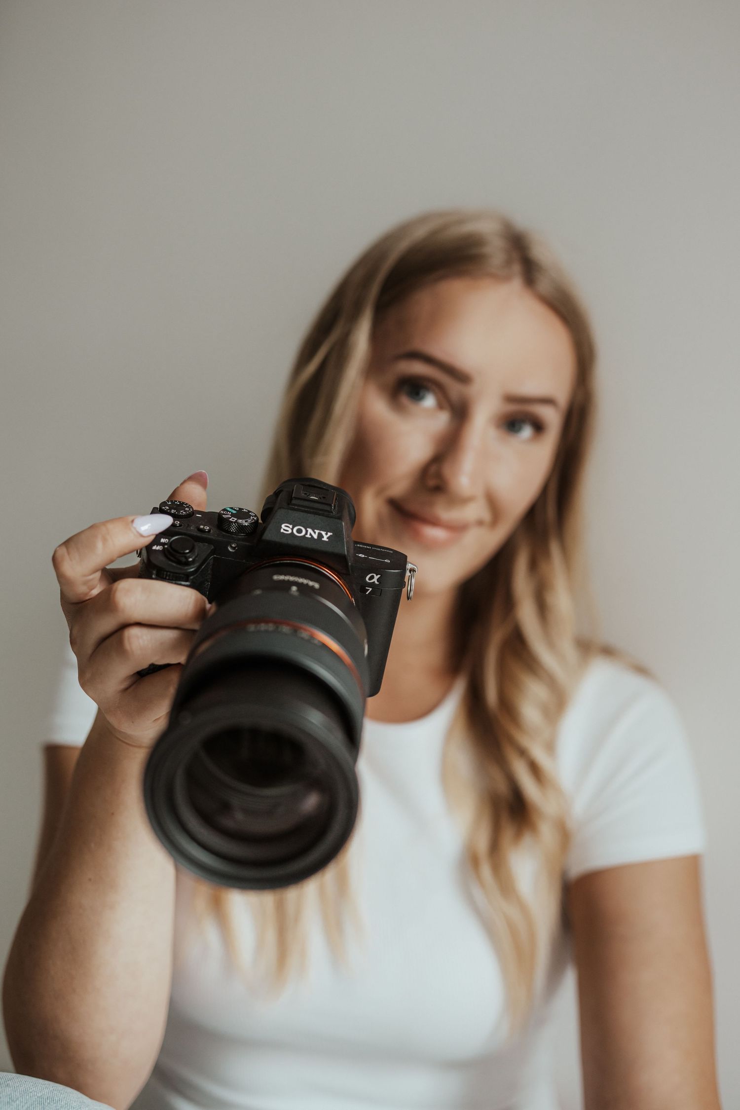 Lächelnde Fotografin im weißen T-Shirt hält eine Sony-Systemkamera mit großem Objektiv in Richtung Kamera. Sarah Appel Fotografie