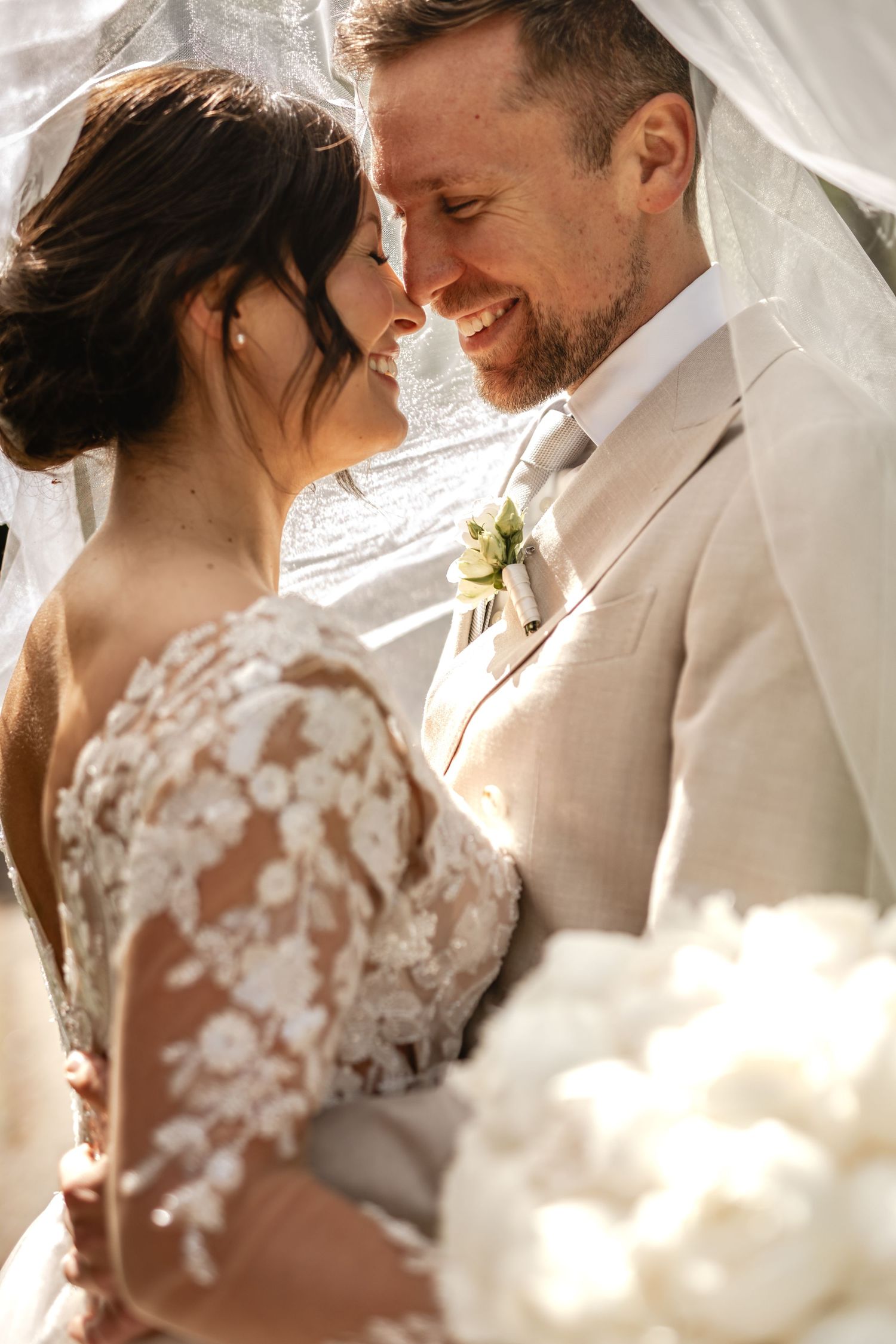 Braut und Bräutigam berühren sich lächelnd mit der Nase unter dem wehenden Hochzeitsschleier. Die Braut hält einen Strauß aus Pfingstrosen.