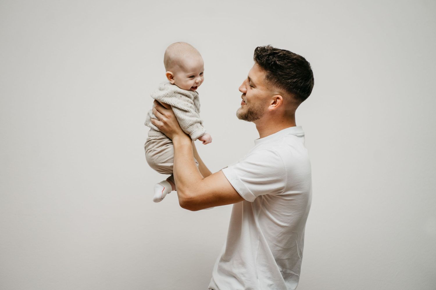 Smiling father in a white t-shirt lifting his baby in the air against a plain light studio background.