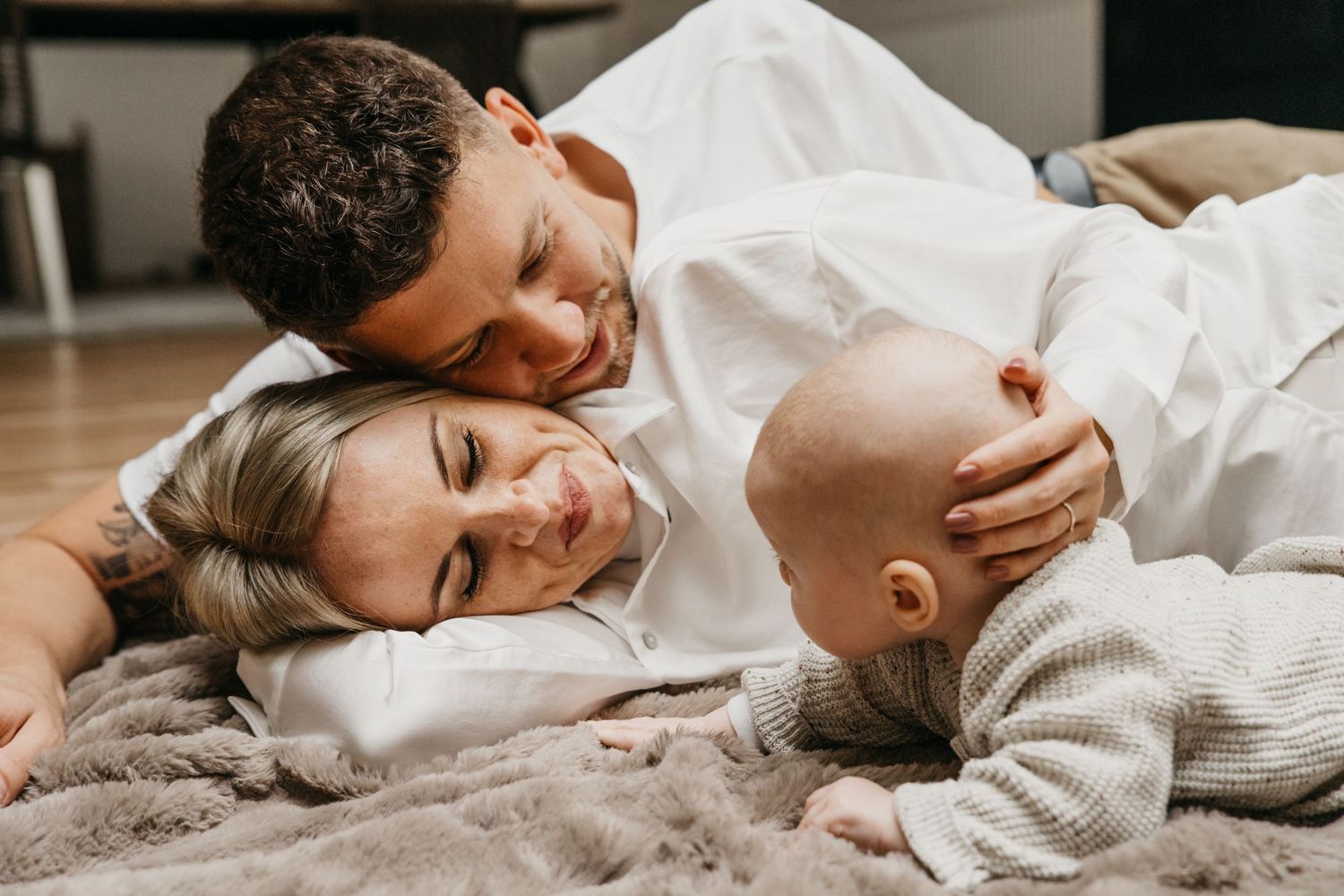 Loving couple in white shirts lying on a soft blanket with their baby, sharing a warm and peaceful family moment.