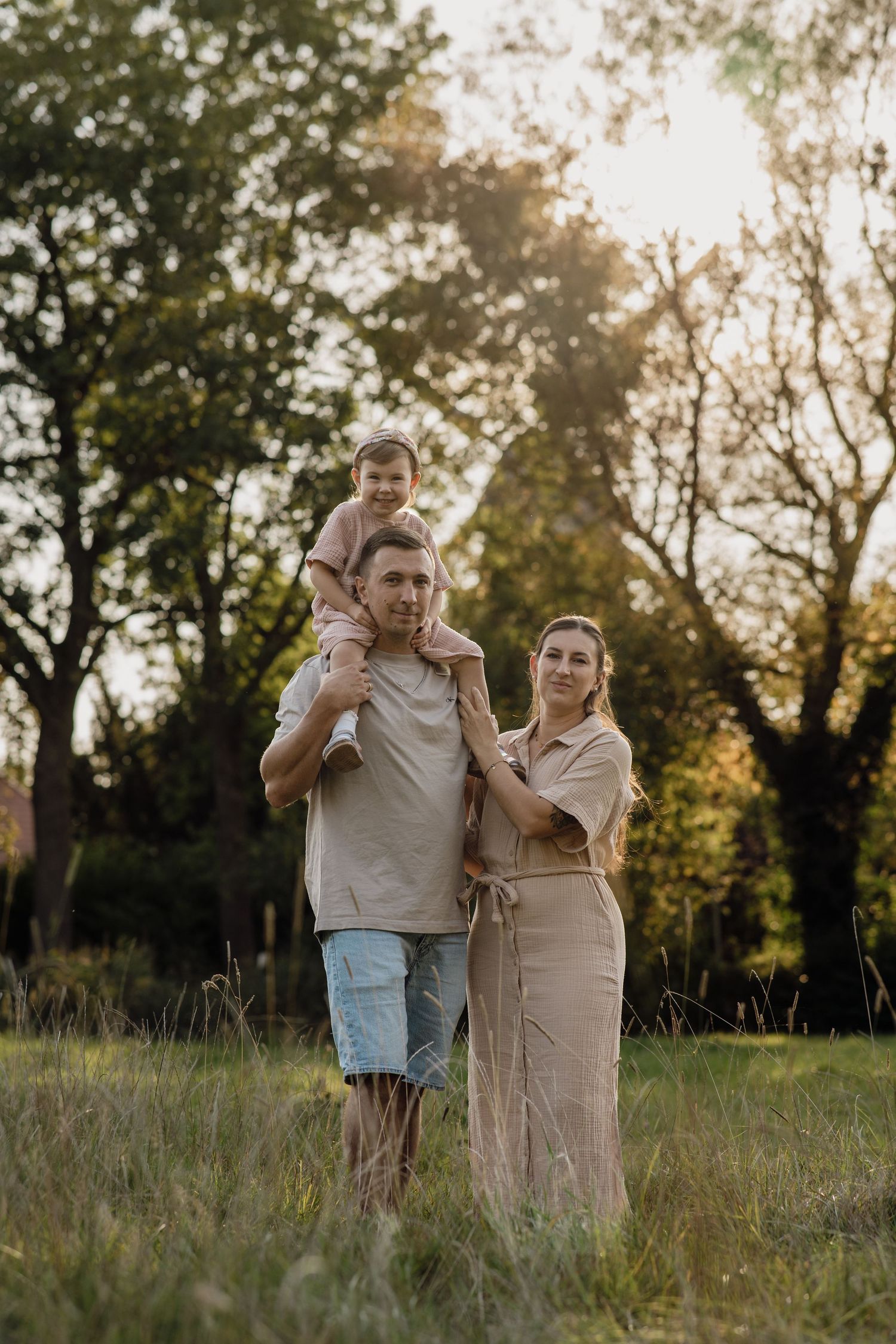 A family poses in a sunlit meadow with a toddler riding on her father's shoulders and the mother standing close beside him.