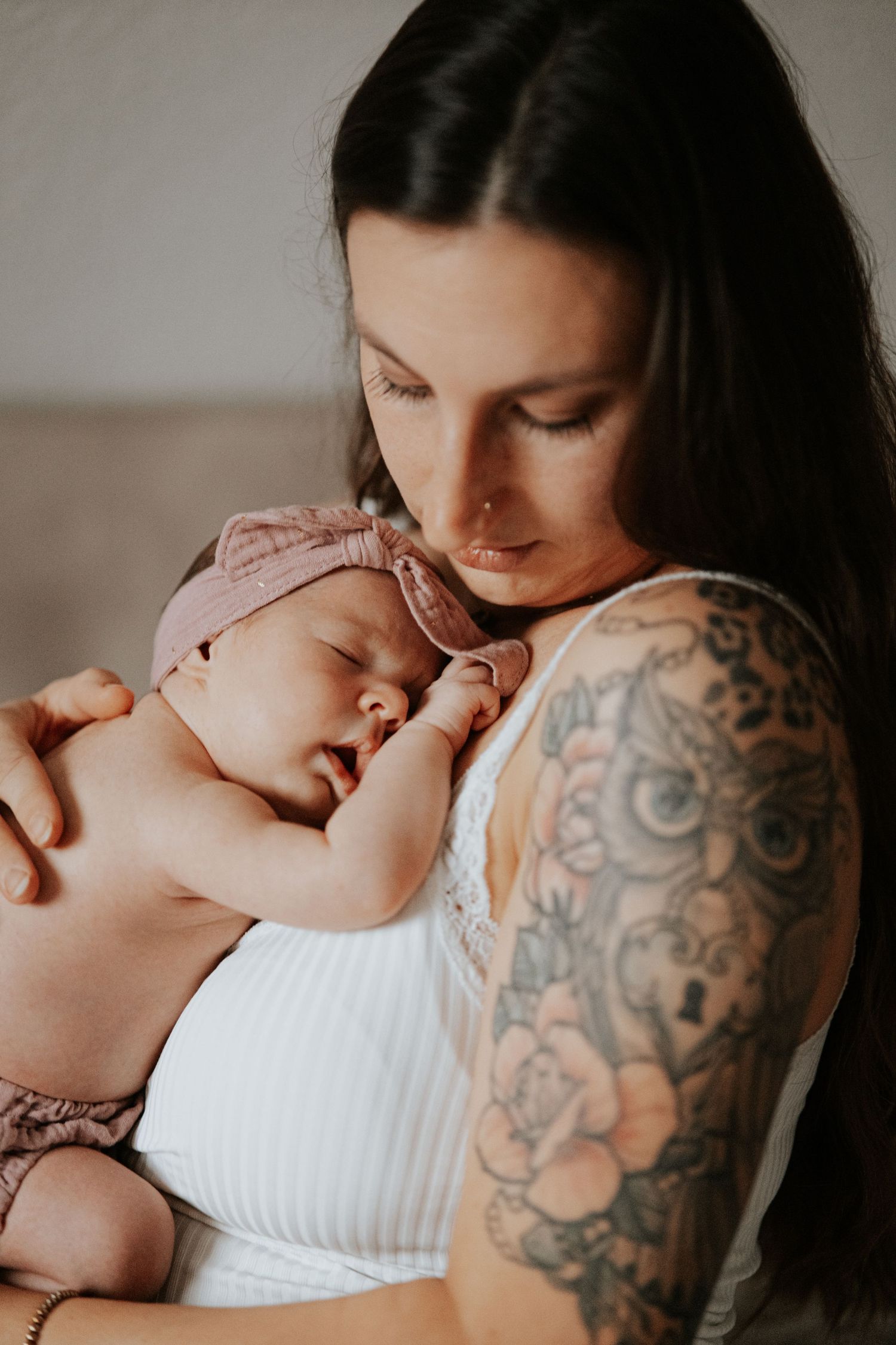 A tattooed mother with long dark hair gazes down at her sleeping newborn wearing a pink knotted headband.
