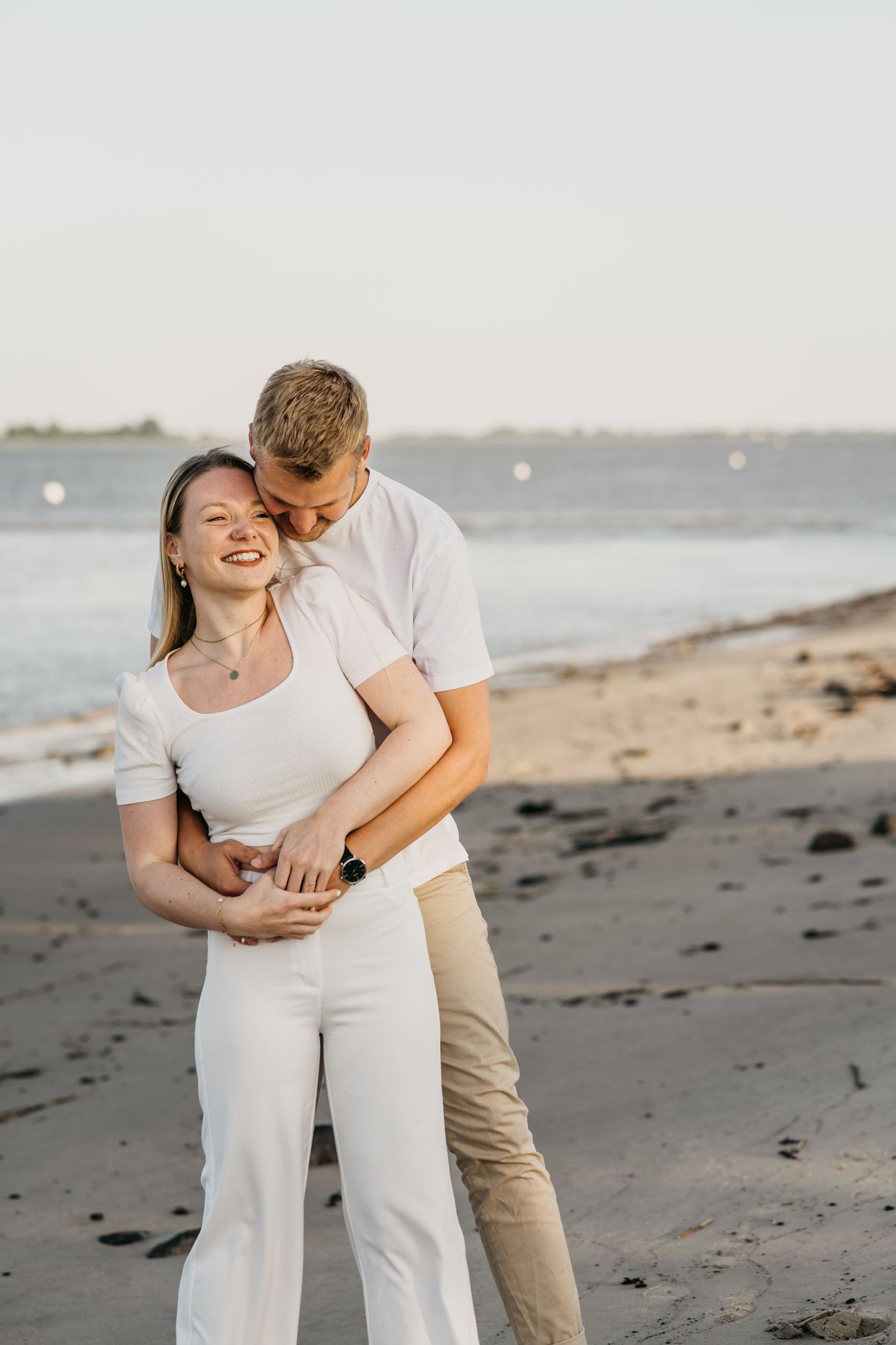 A smiling couple in white outfits embraces on a sandy beach at sunset with calm water in the background.