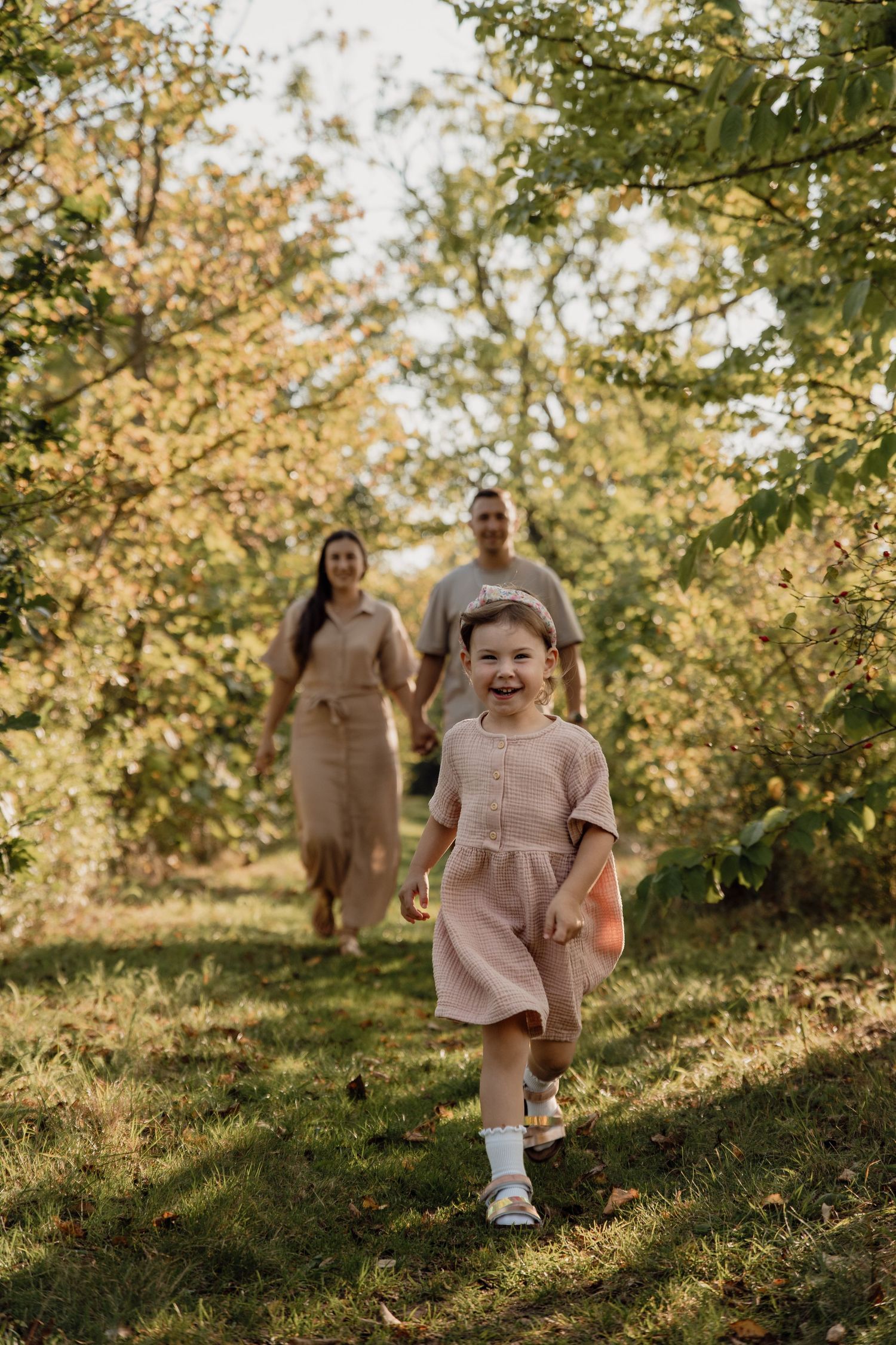 A laughing little girl in a pink dress runs through a sunlit orchard while her parents walk hand in hand behind her.