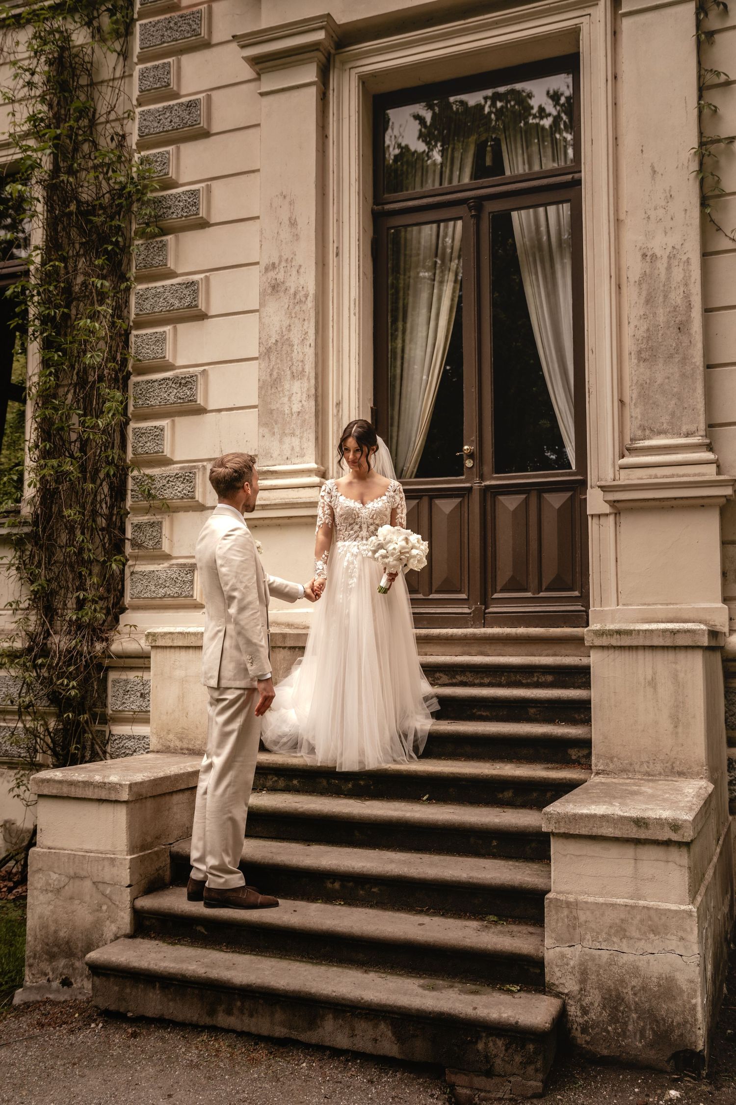 Braut und Bräutigam stehen Hand in Hand auf einer Steintreppe vor einer historischen Villa mit hohen Holztüren und efeubewachsenen Mauern.