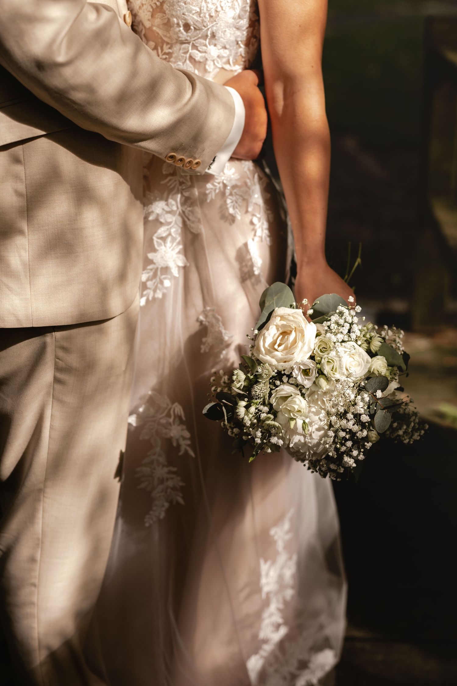 Bräutigam in beigem Anzug umarmt seine Braut im Spitzenkleid. Sie hält einen Brautstrauß aus weißen Rosen und Schleierkraut. Neuenwalde