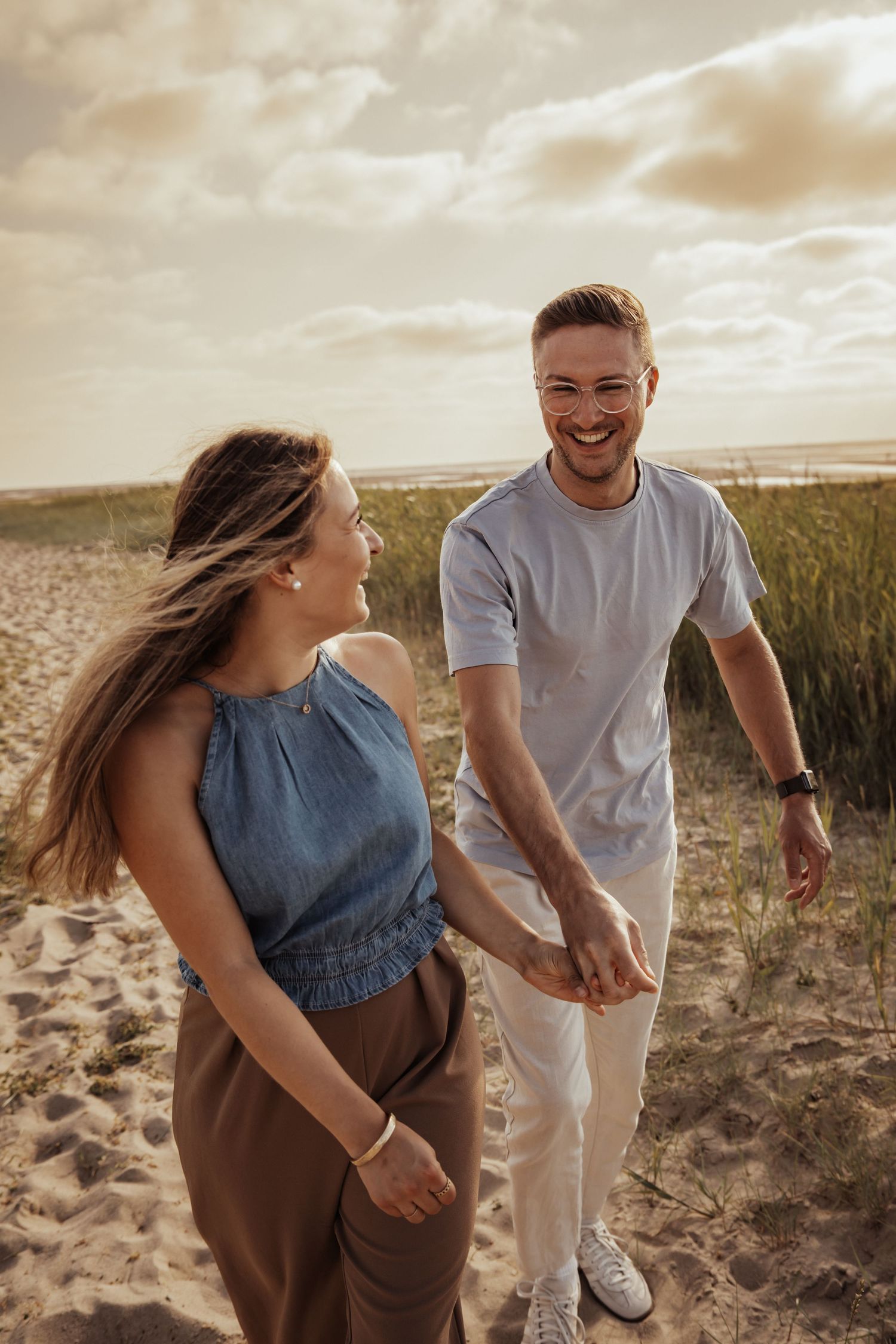 A laughing couple holds hands and walks along a sandy beach path with windswept hair under a soft golden sunset sky.