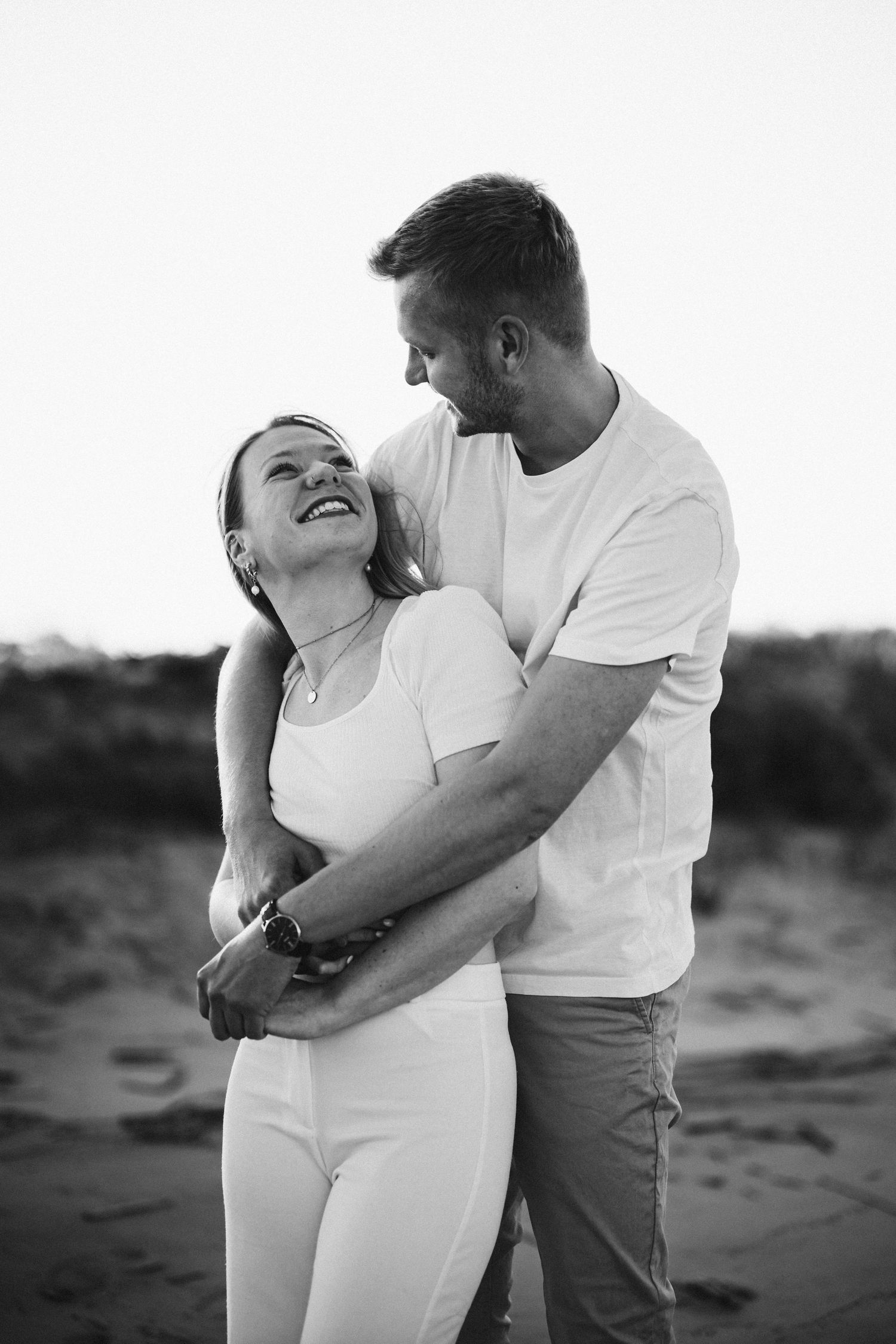 Black and white photo of a laughing woman looking up at her partner as he hugs her from behind on a beach.