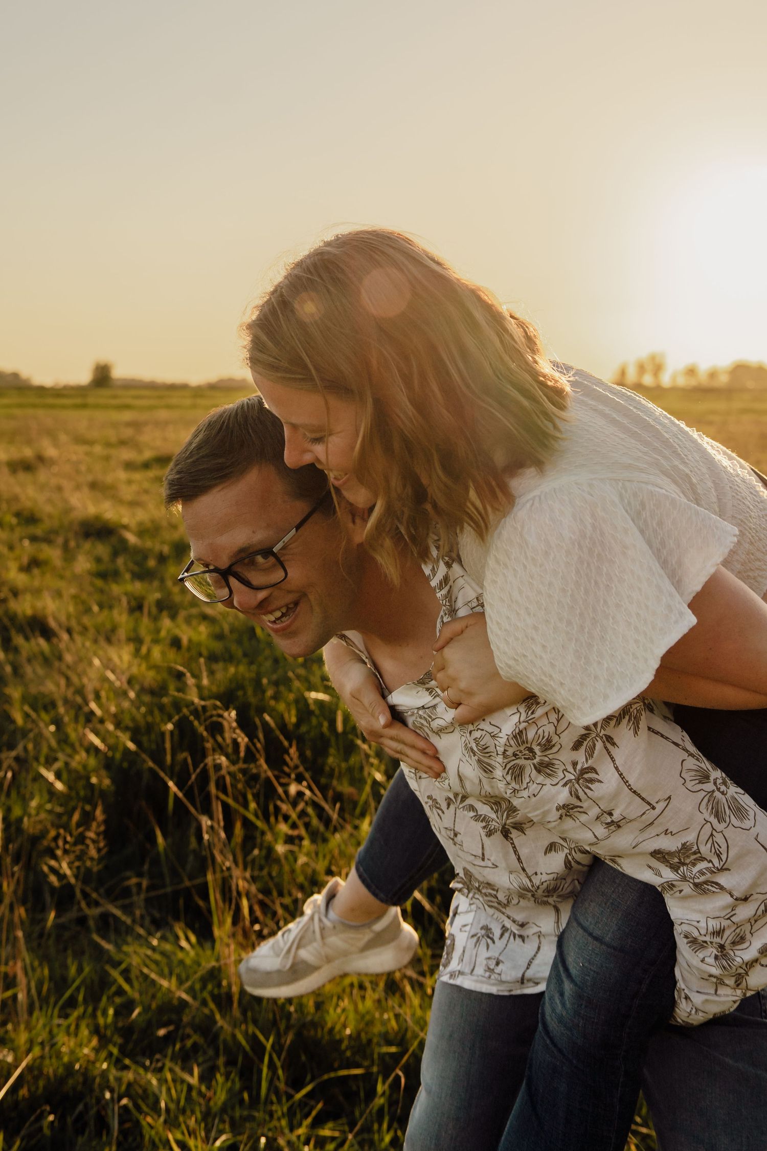 Laughing man in glasses gives his partner a piggyback ride across a grassy field at sunset.