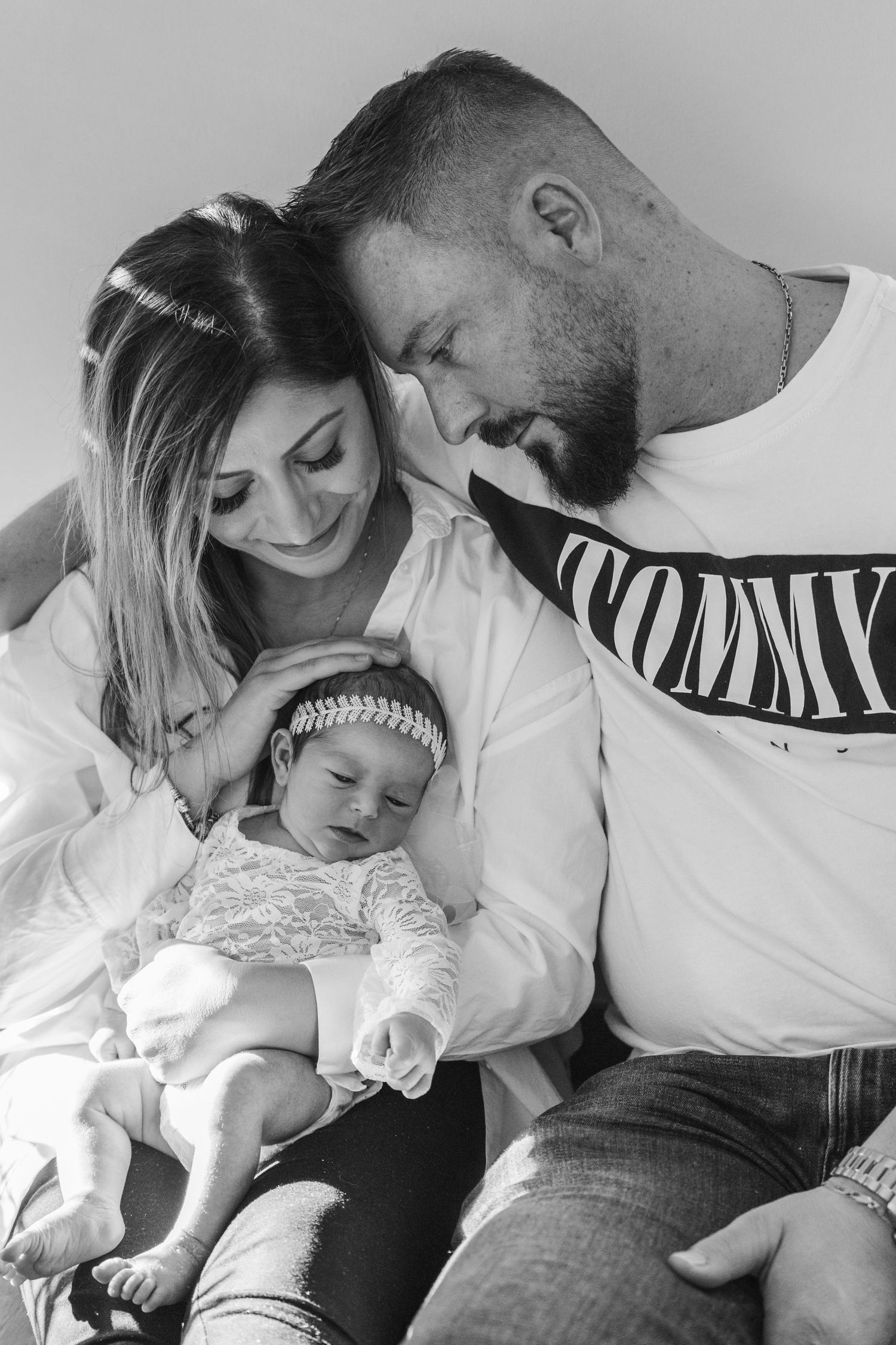 Black and white portrait of a mother and father gently holding their sleeping newborn baby in a lace outfit and headband.