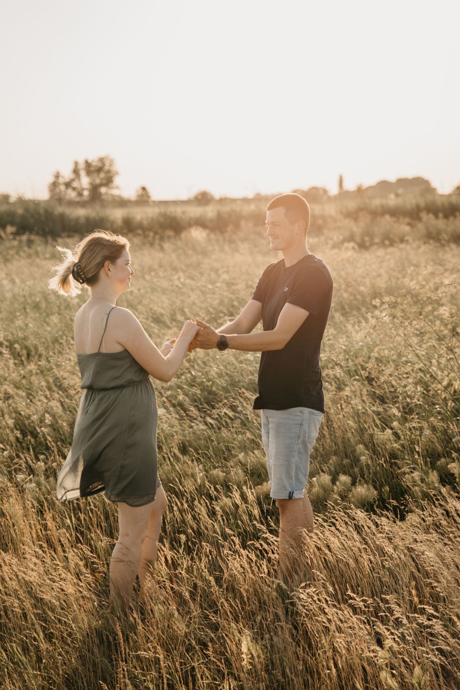 Couple holding hands and facing each other in a tall grass field bathed in warm golden hour sunlight.