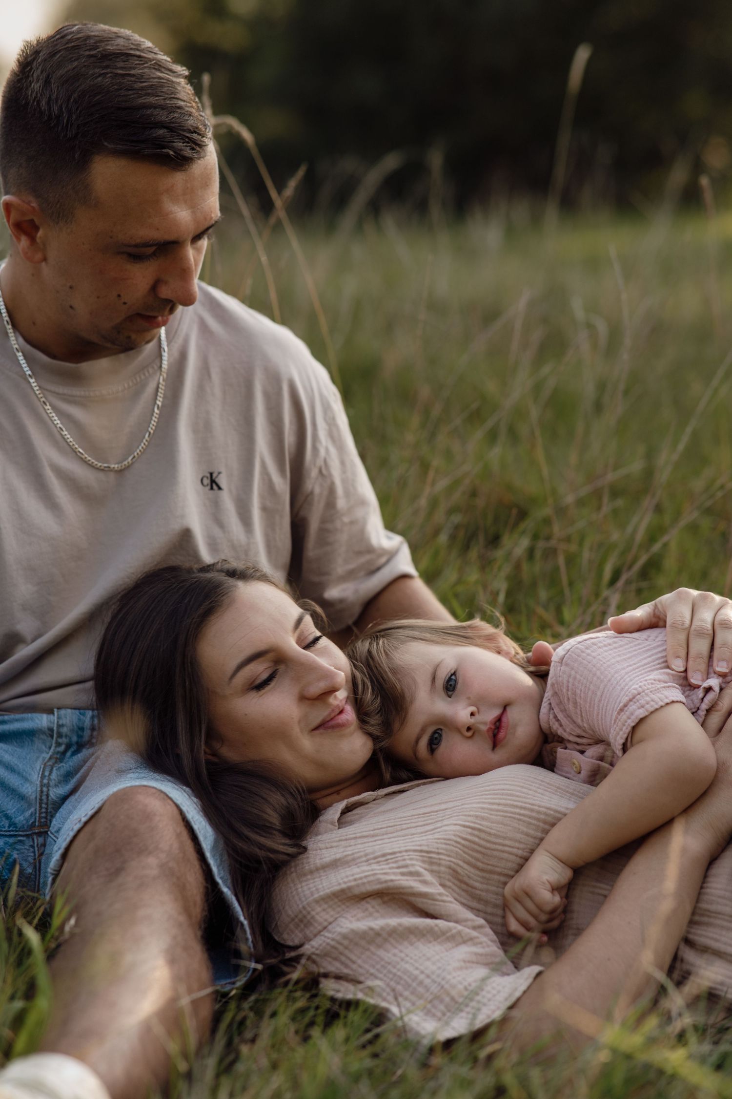A father and mother relax in the grass, cuddling their young daughter during a warm outdoor family portrait session.