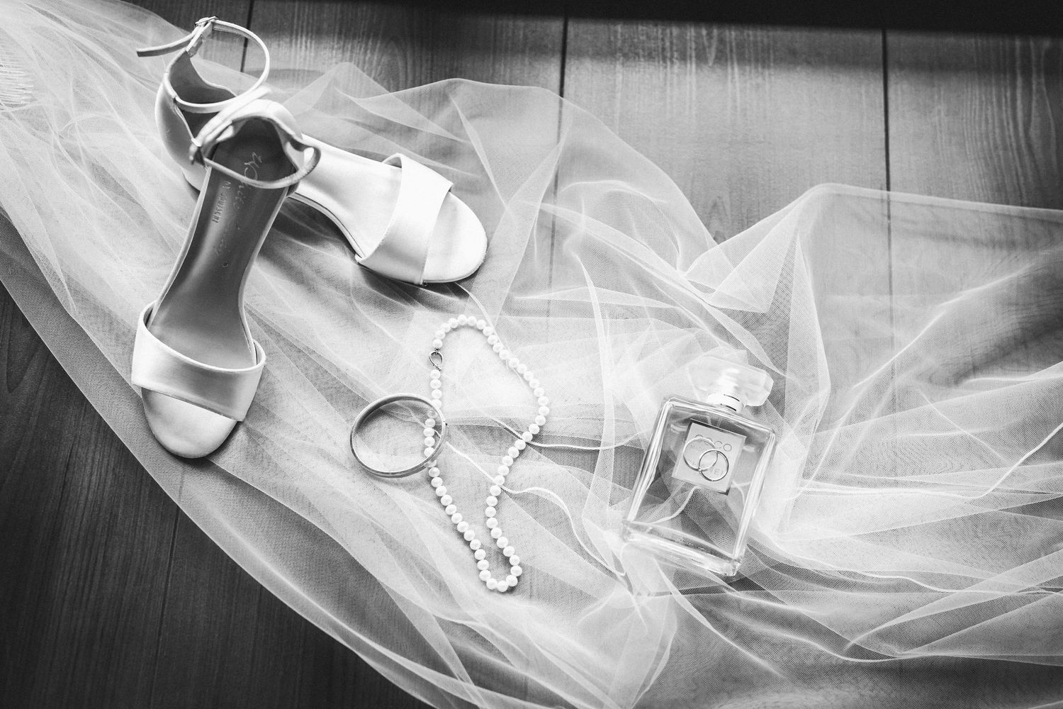Schwarz-Weiß-Detailaufnahme von Brautschuhen mit Absatz. Perlenkette, Armreif und Parfümflakon auf fließendem Tüllstoff.