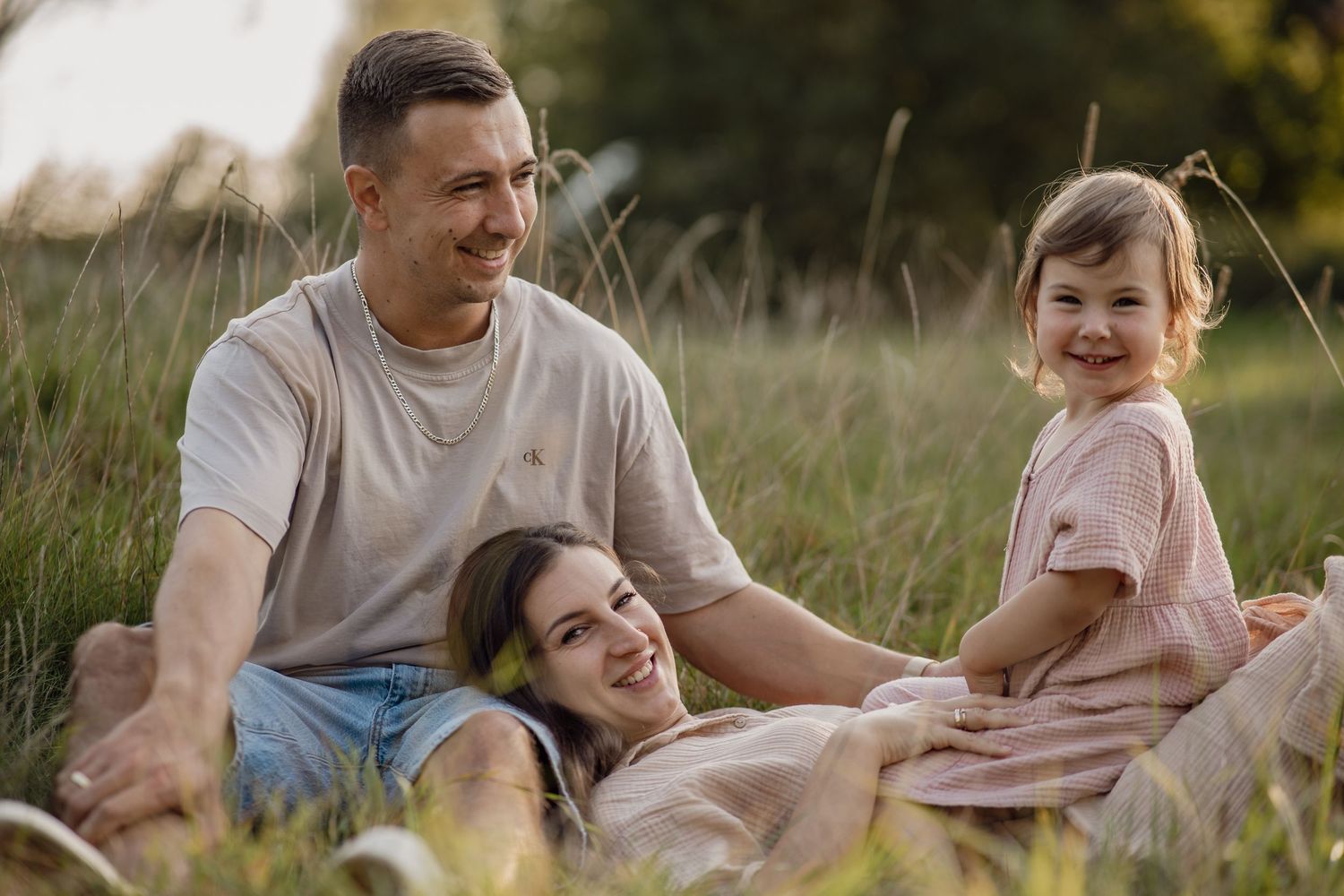 Familie mit Vater, Mutter und kleiner Tochter liegt entspannt im grünen Gras und genießt gemeinsam den Sonnenuntergang.