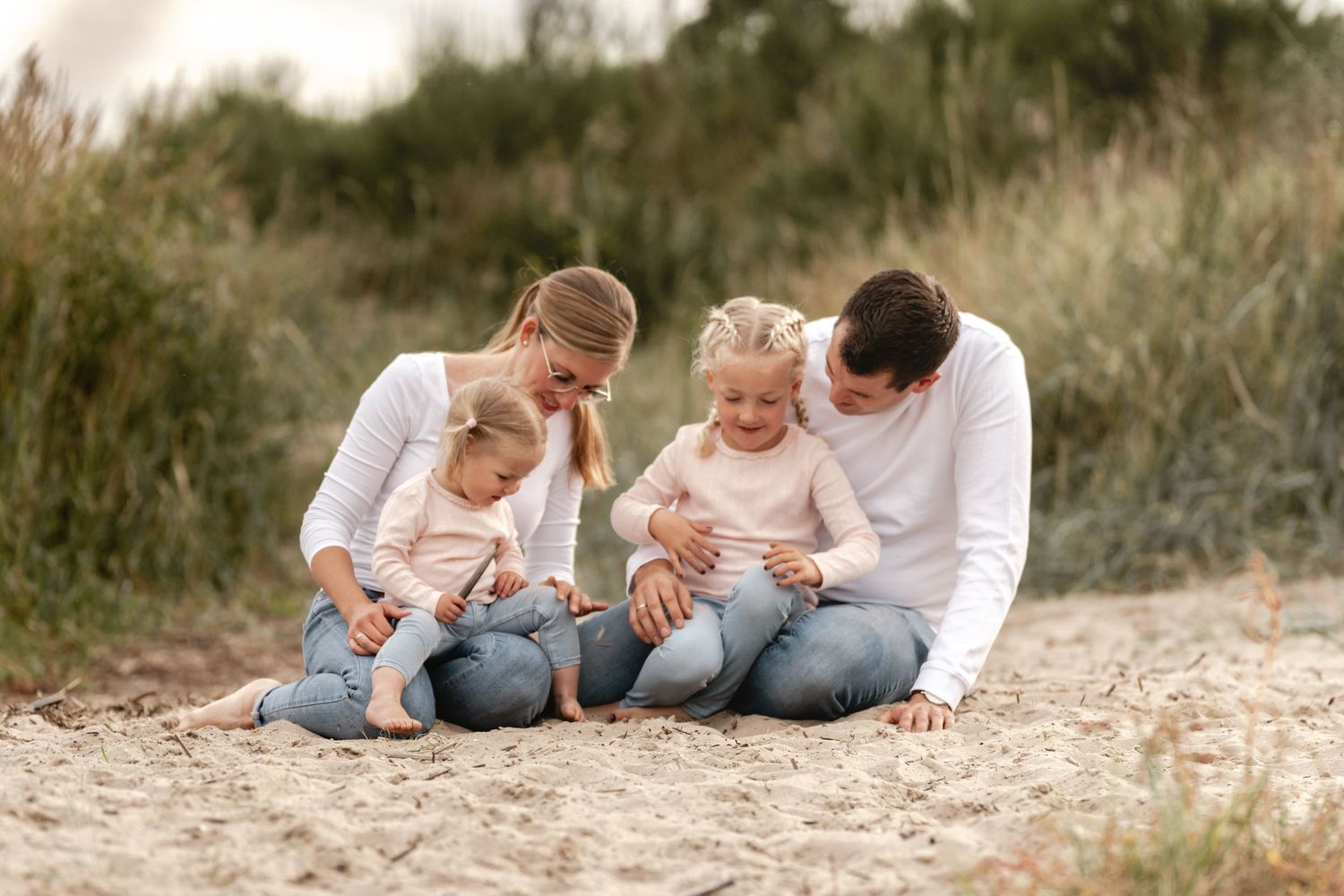 Family of four with two young daughters sitting together on a sandy dune, dressed in white and soft neutral tones.