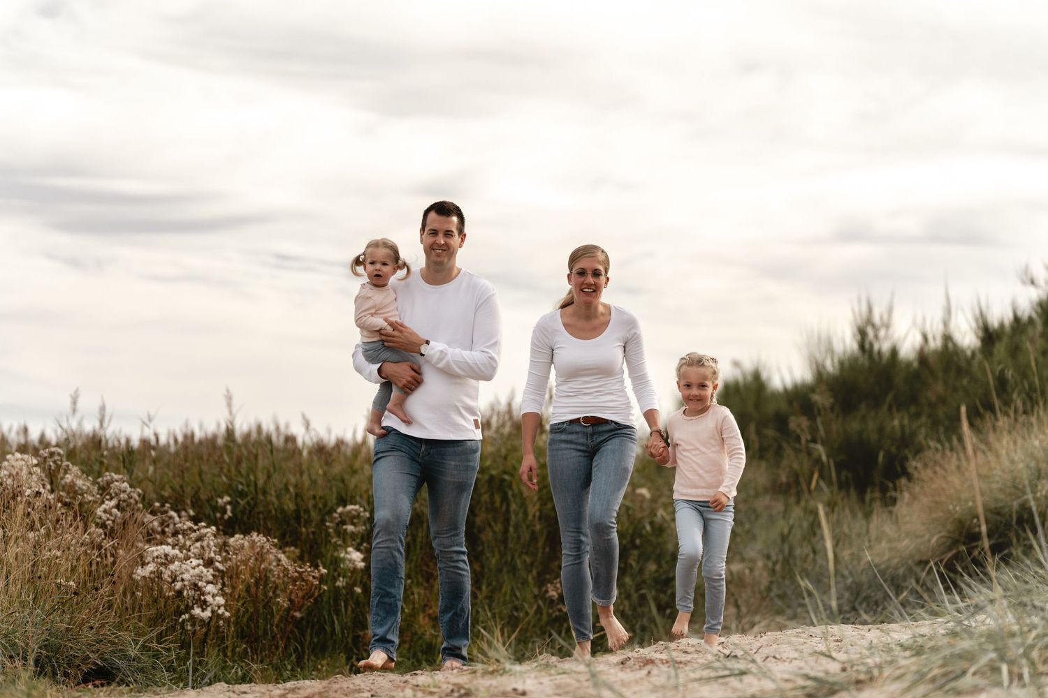 A smiling family of four walks along a sandy dune path with tall beach grass under a soft cloudy sky.