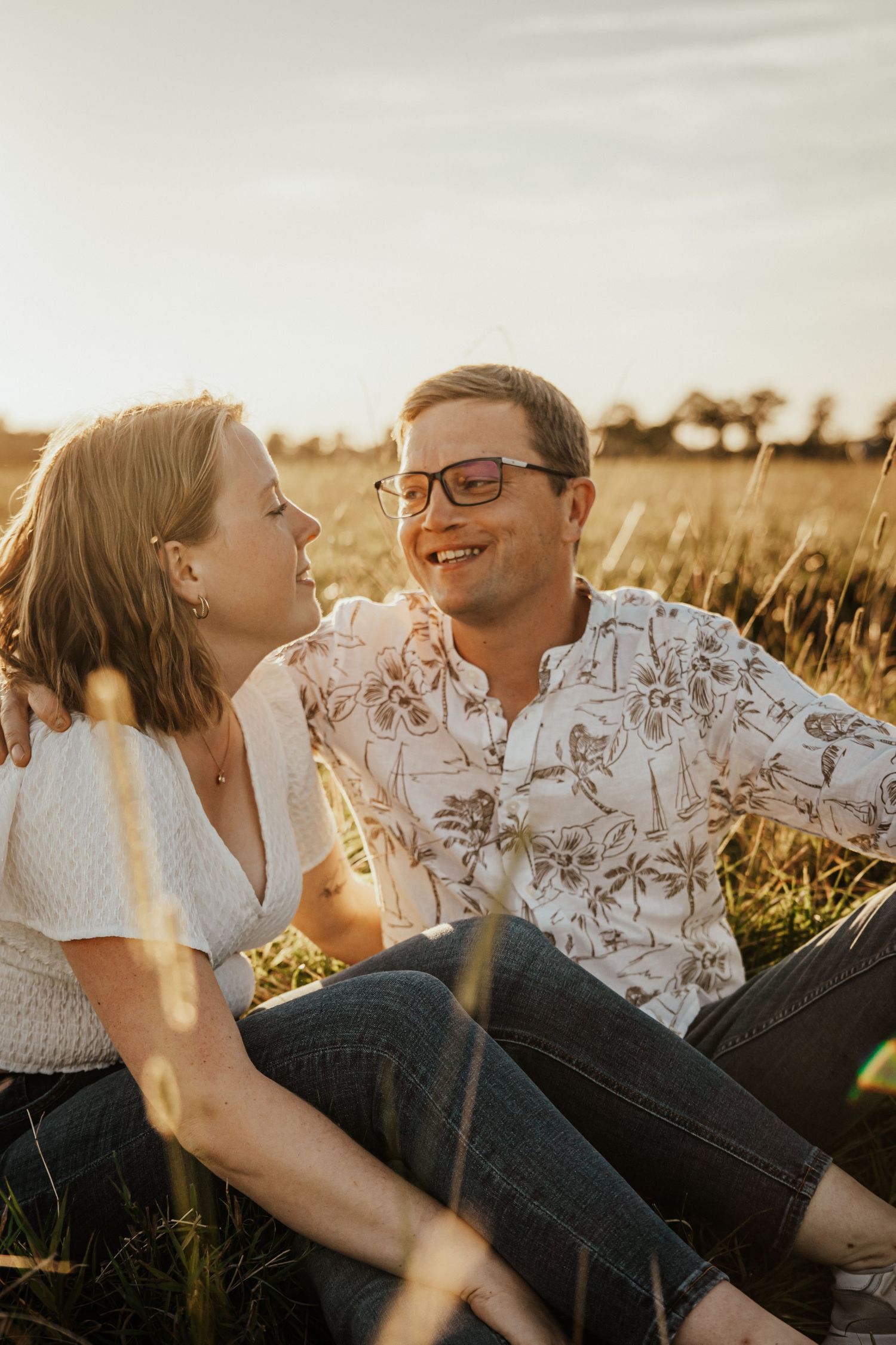 Smiling couple sitting close together in a golden grass field at sunset, the woman leaning in toward the man in glasses.