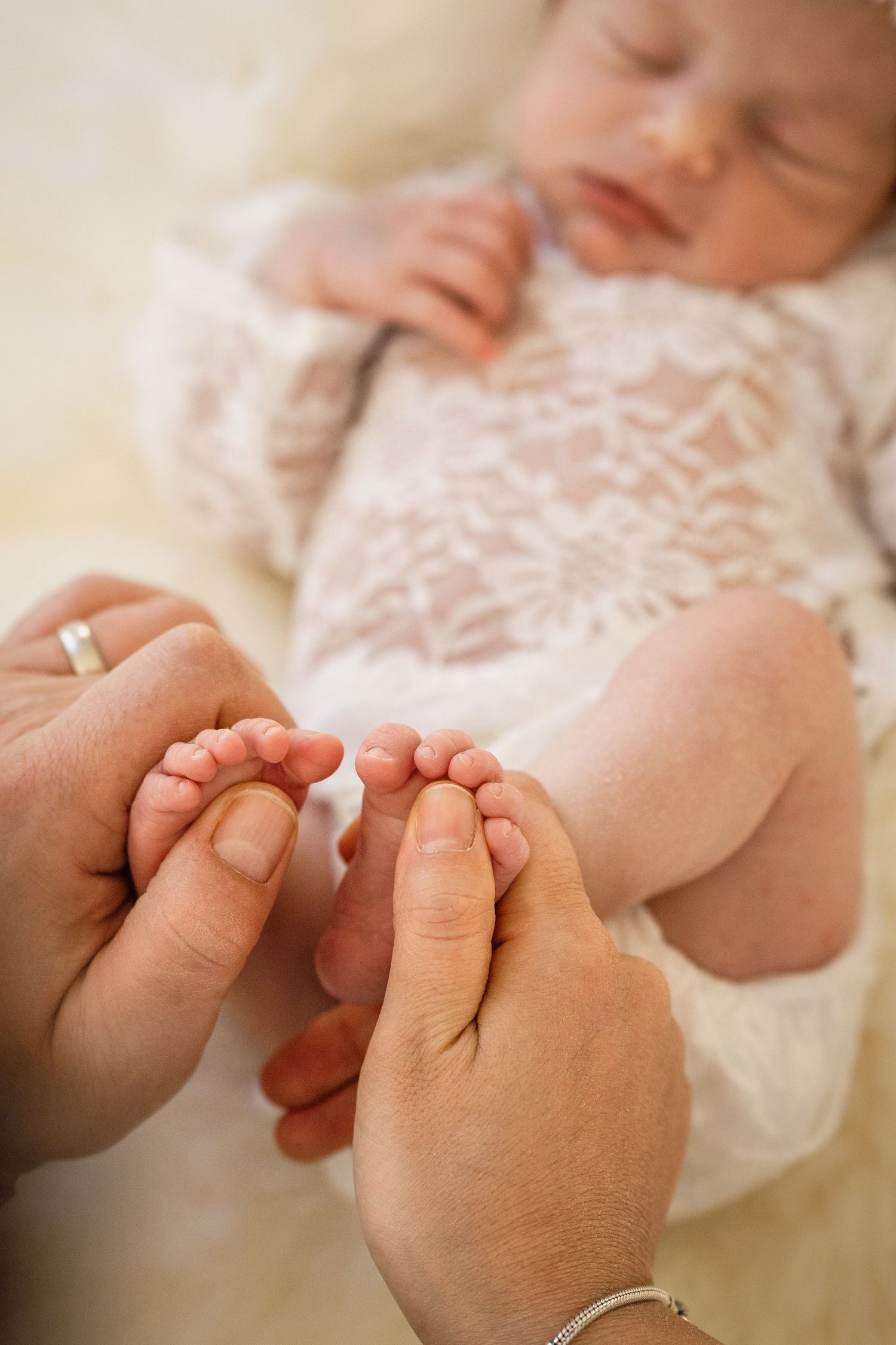 Parents gently holding a newborn baby's tiny feet while the baby sleeps in a delicate white lace outfit.