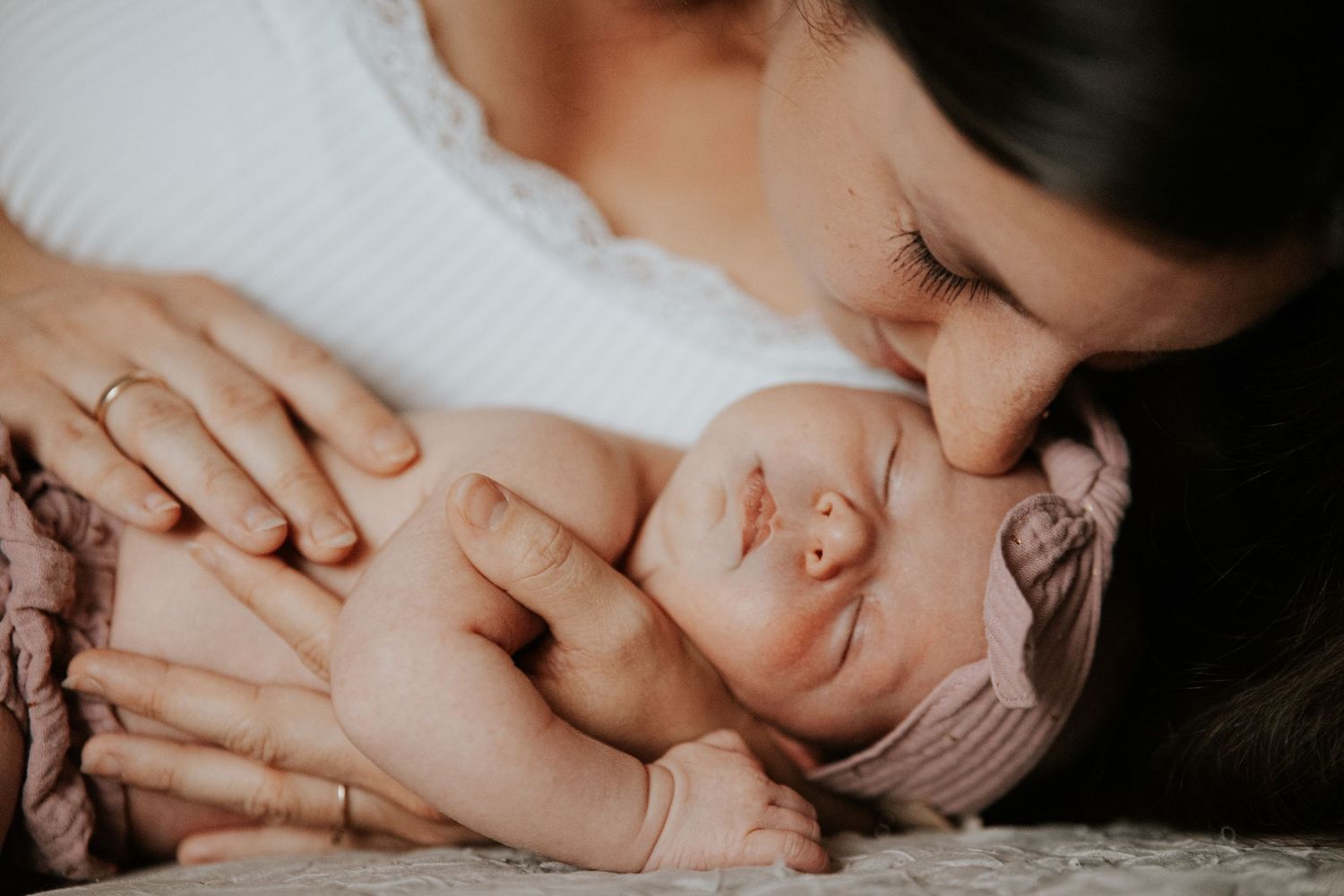 Nahaufnahme einer Mutter, die ihr schlafendes Neugeborenes mit rosa Haarband liebevoll küsst und in den Armen hält.
