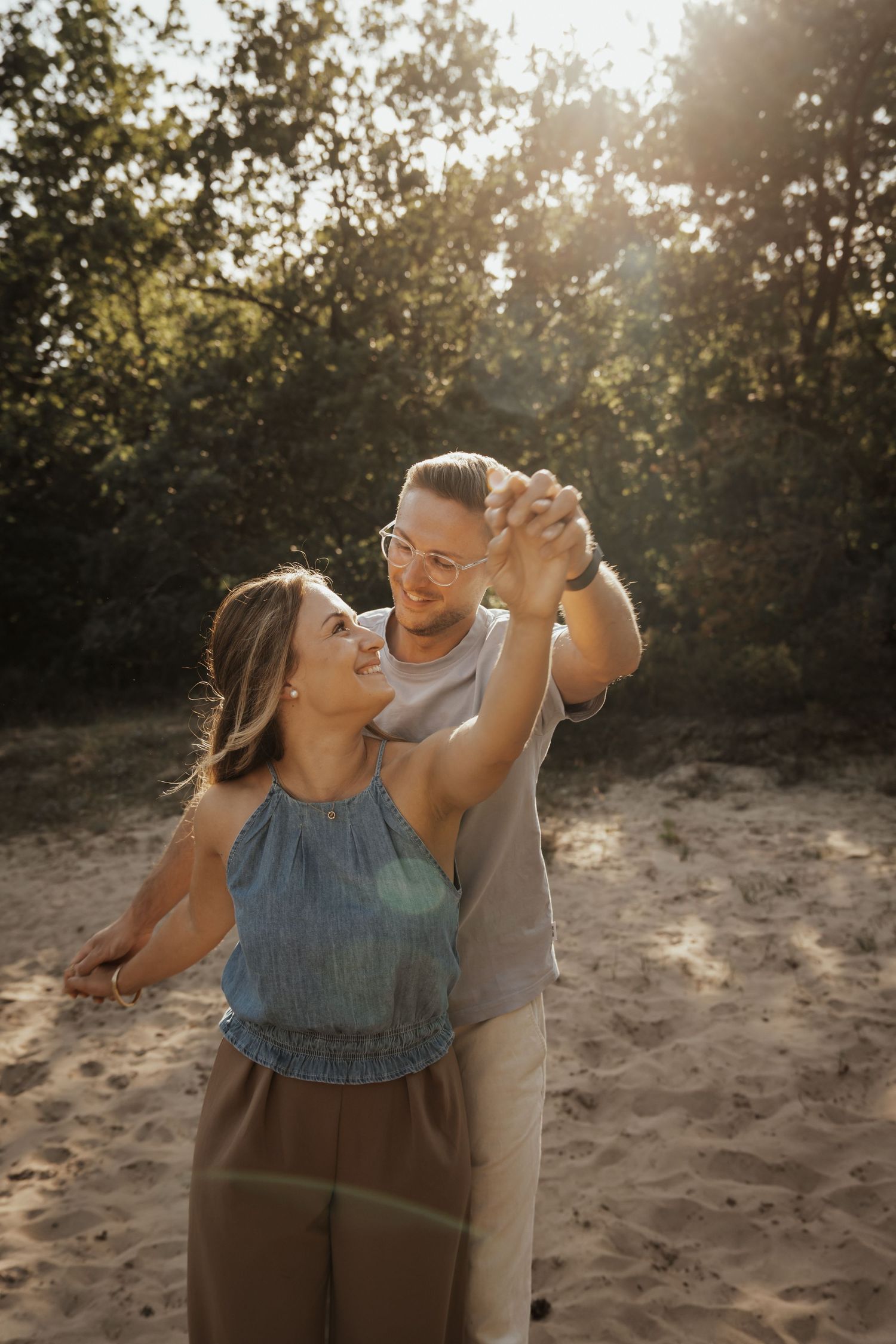 A cheerful couple dances hand in hand on a sandy woodland clearing, glowing in the soft light of the afternoon sun.