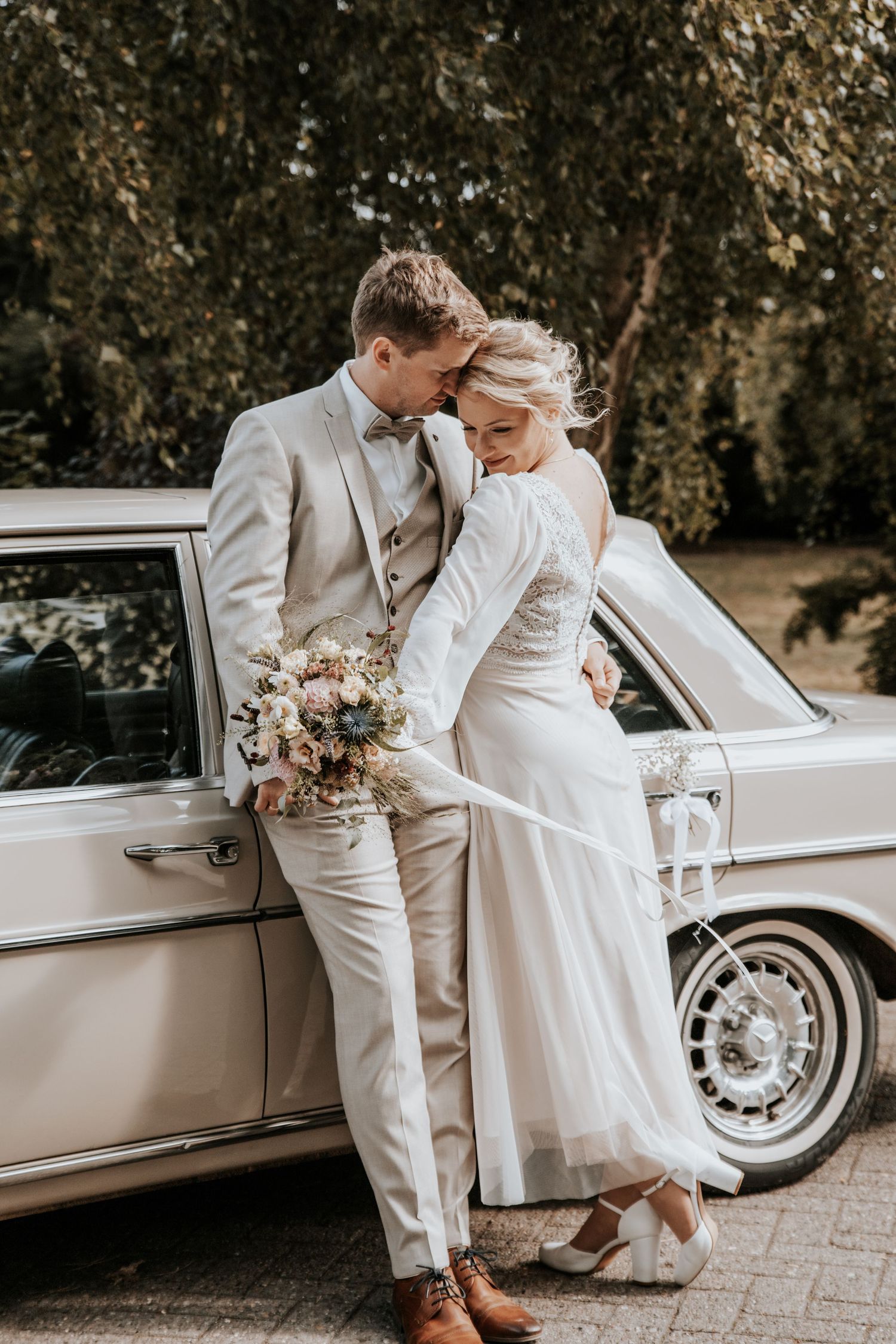 Braut im langärmligen Spitzenkleid und Bräutigam im beigen Anzug umarmen sich neben einem cremefarbenen Mercedes Oldtimer. Weserterrassen