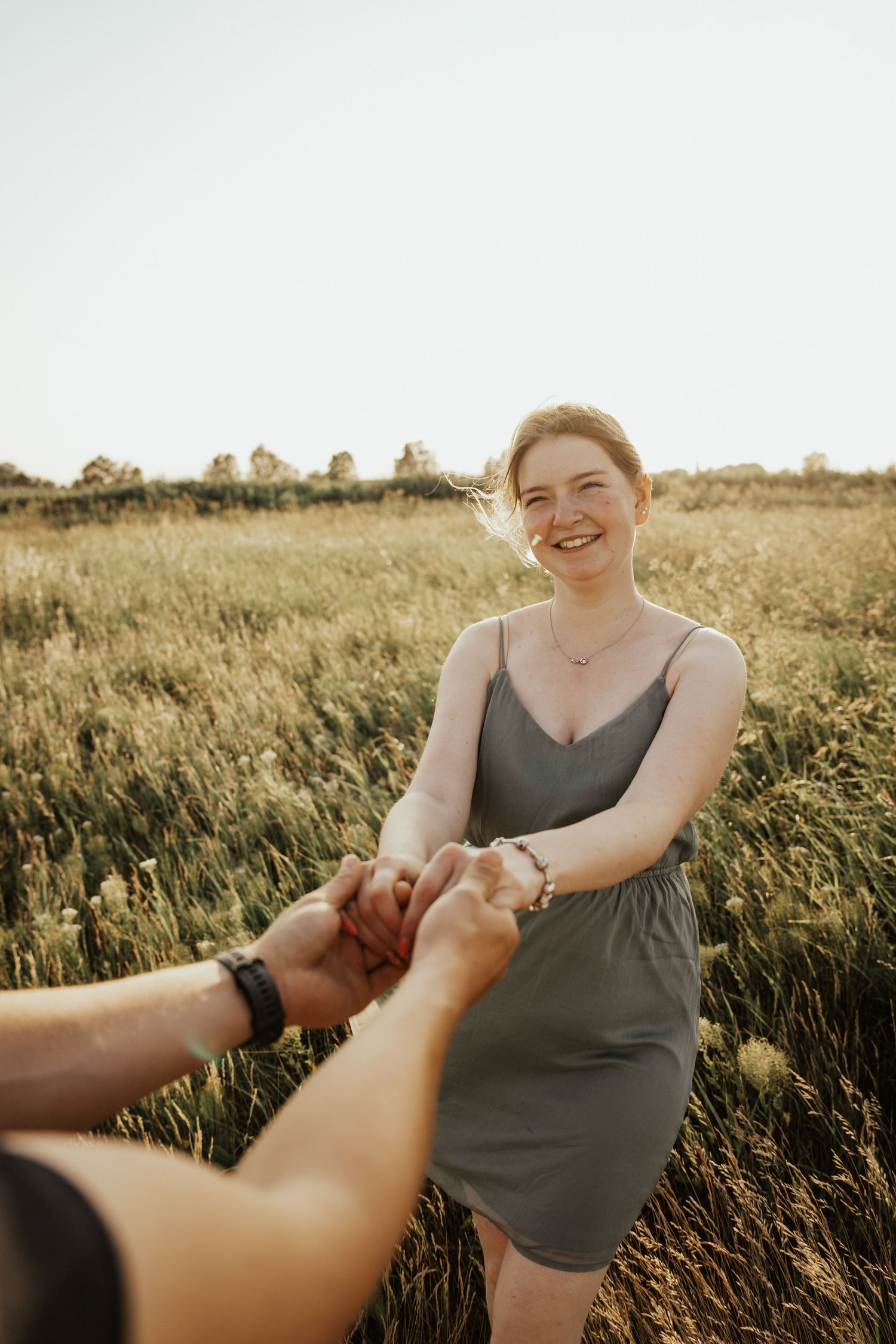 Smiling woman in a green dress holds hands with her partner in a sunny field of tall golden grass.