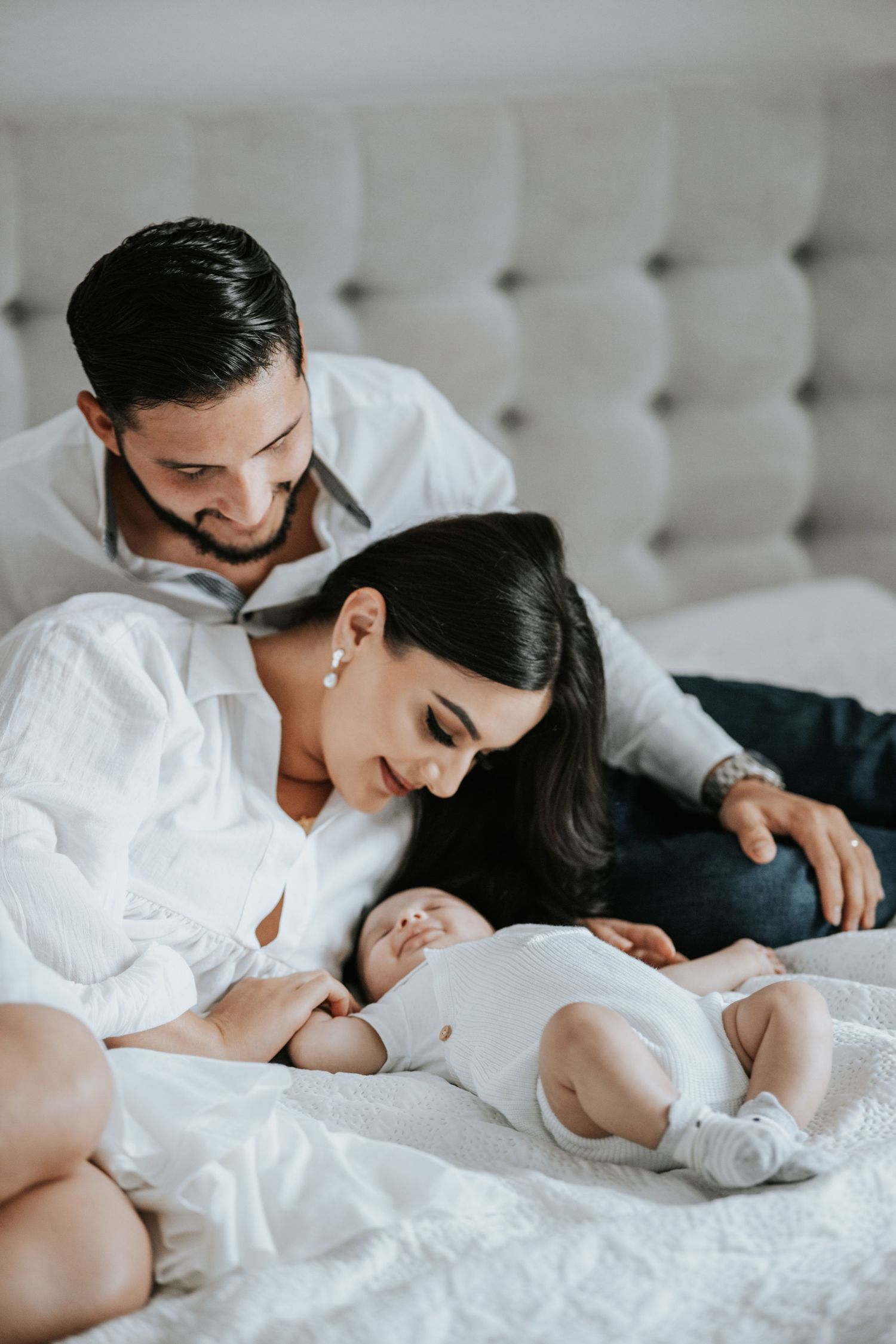 Smiling mother and father in white outfits cuddle their newborn baby lying on a bed in a bright bedroom.