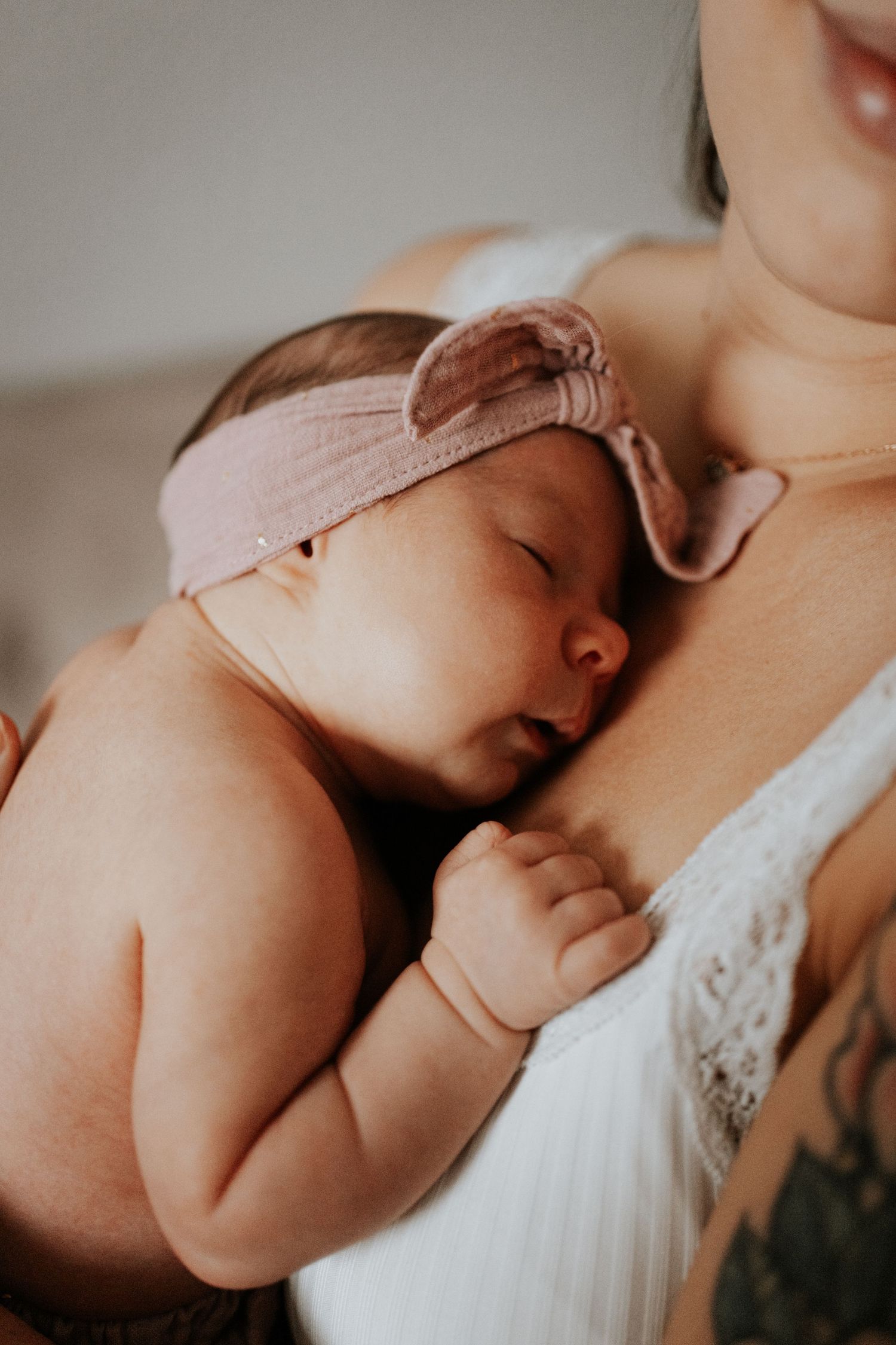 Schlafendes Neugeborenes mit rosa Haarband liegt an der Brust seiner Mutter und hält mit einer kleinen Hand ihr Spitzentop fest.