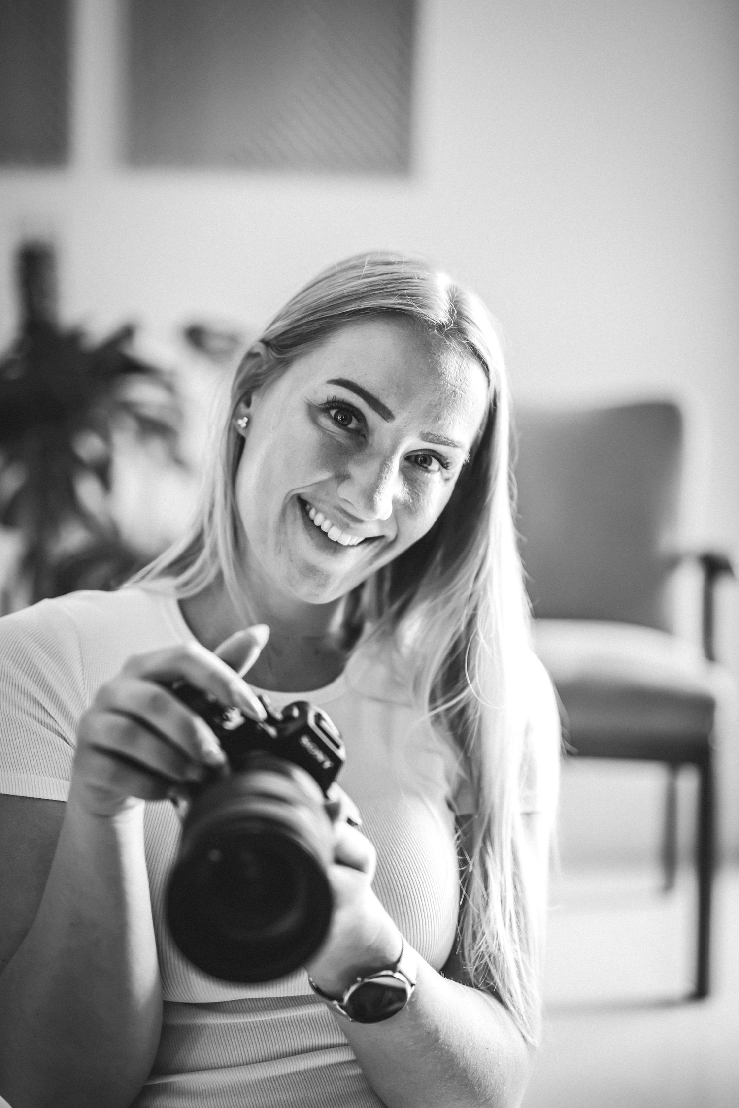 Schwarz-Weiß-Porträt einer lächelnden Fotografin mit langen Haaren, die in einem hellen Raum ihre Kamera hält. Sarah Appel Fotografie