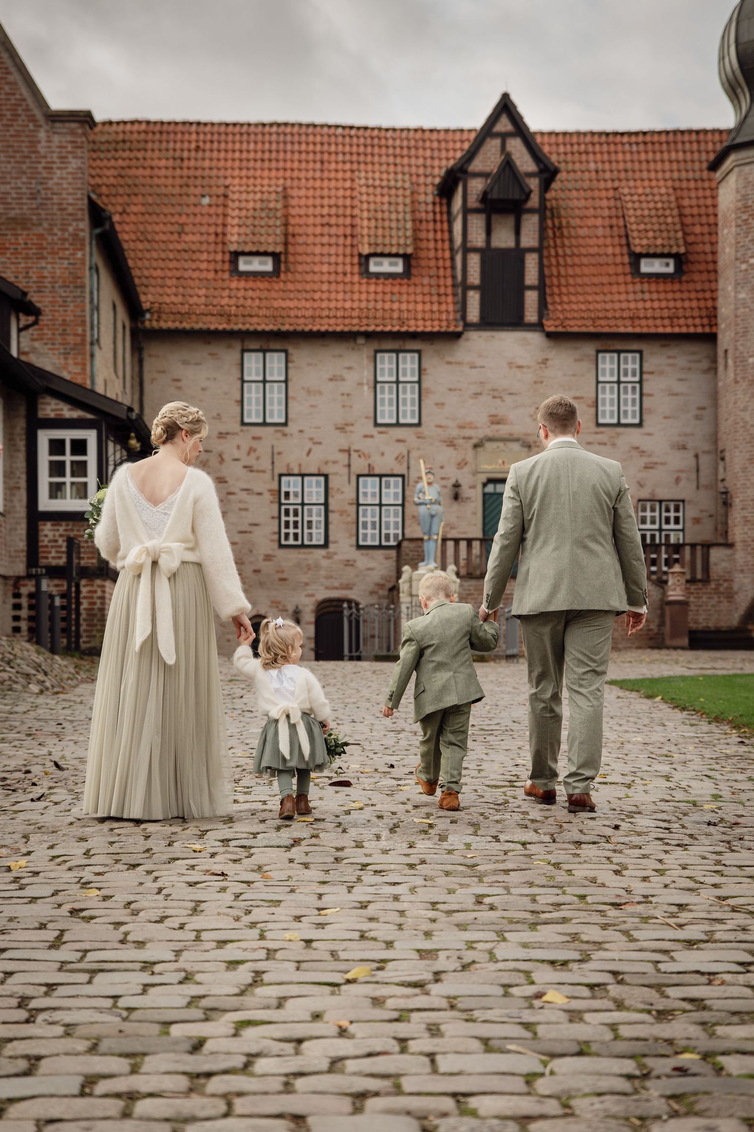 Brautpaar in salbeigrüner Hochzeitskleidung geht Hand in Hand mit Kindern über einen gepflasterten Innenhof auf eine Burg zu. Bad Bederkesa