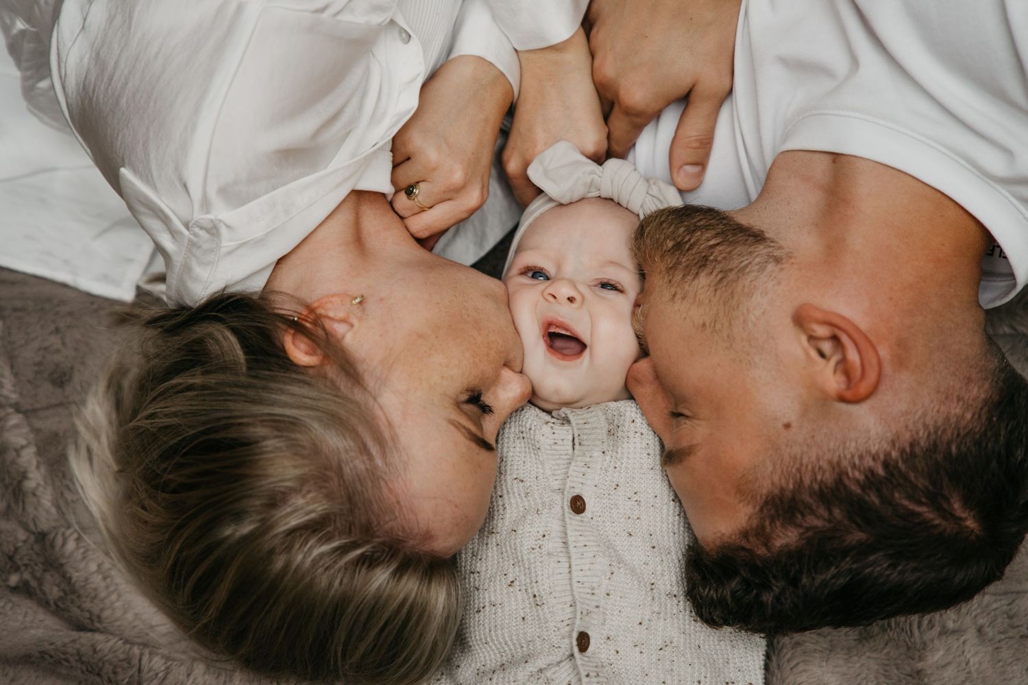 Neugeborenes Baby lächelt zwischen seinen Eltern, die ihm liebevoll einen Kuss auf die Wangen geben.