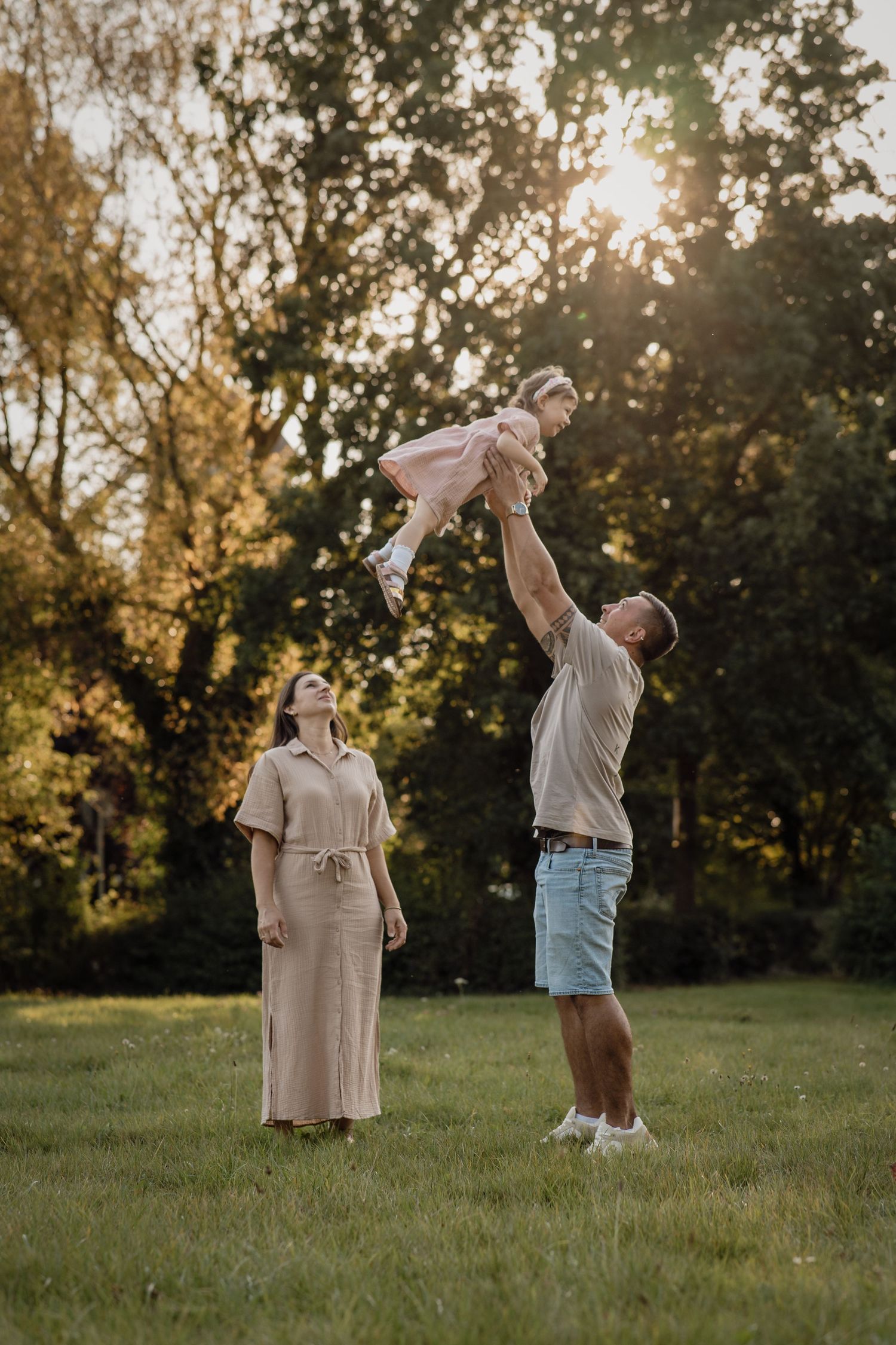 A father lifts his giggling daughter high into the air while the mother watches in a sunny grassy park at sunset.