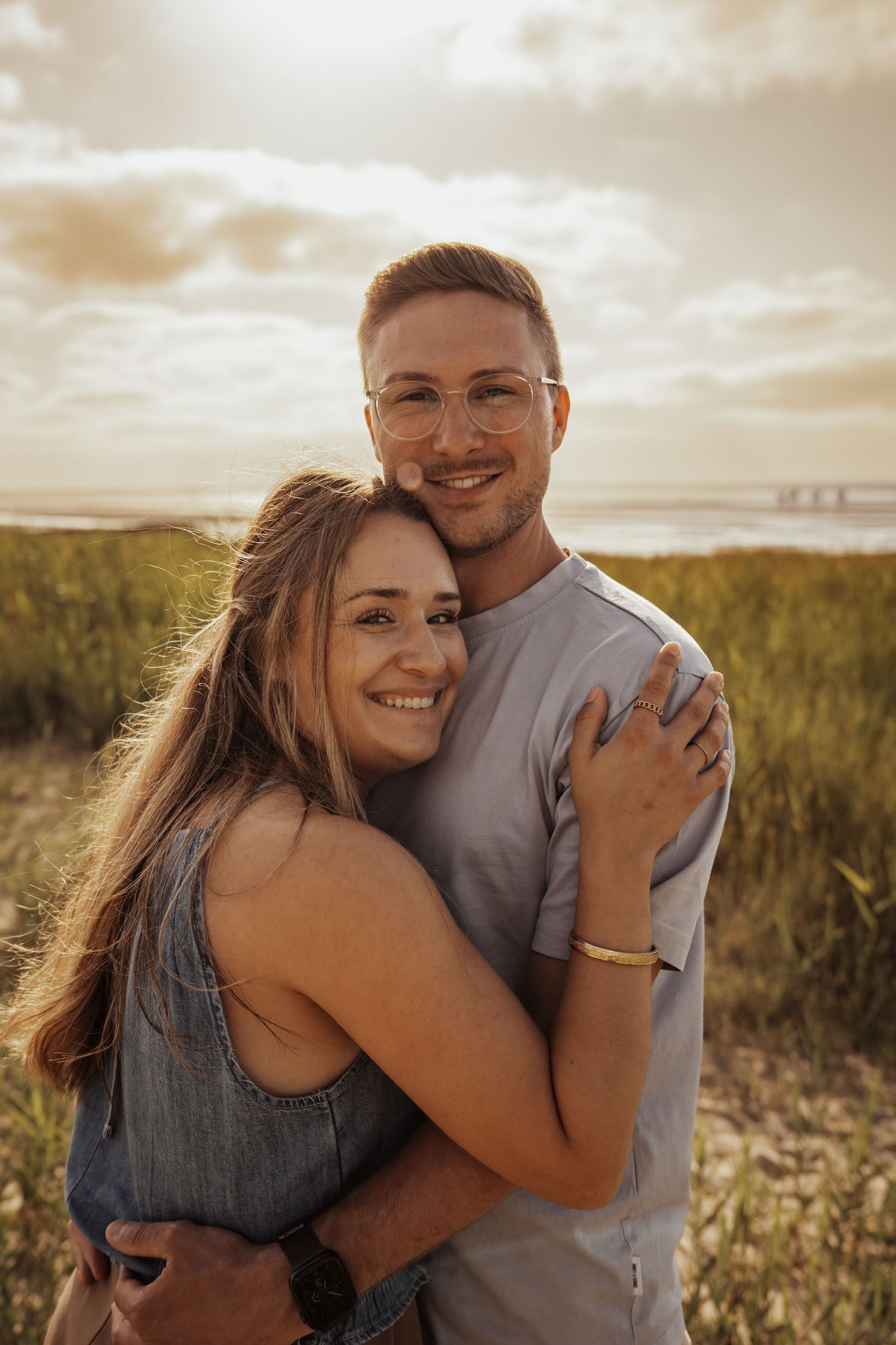 A happy couple embraces and smiles on a coastal dune at golden hour with dune grass and cloudy sky behind them.