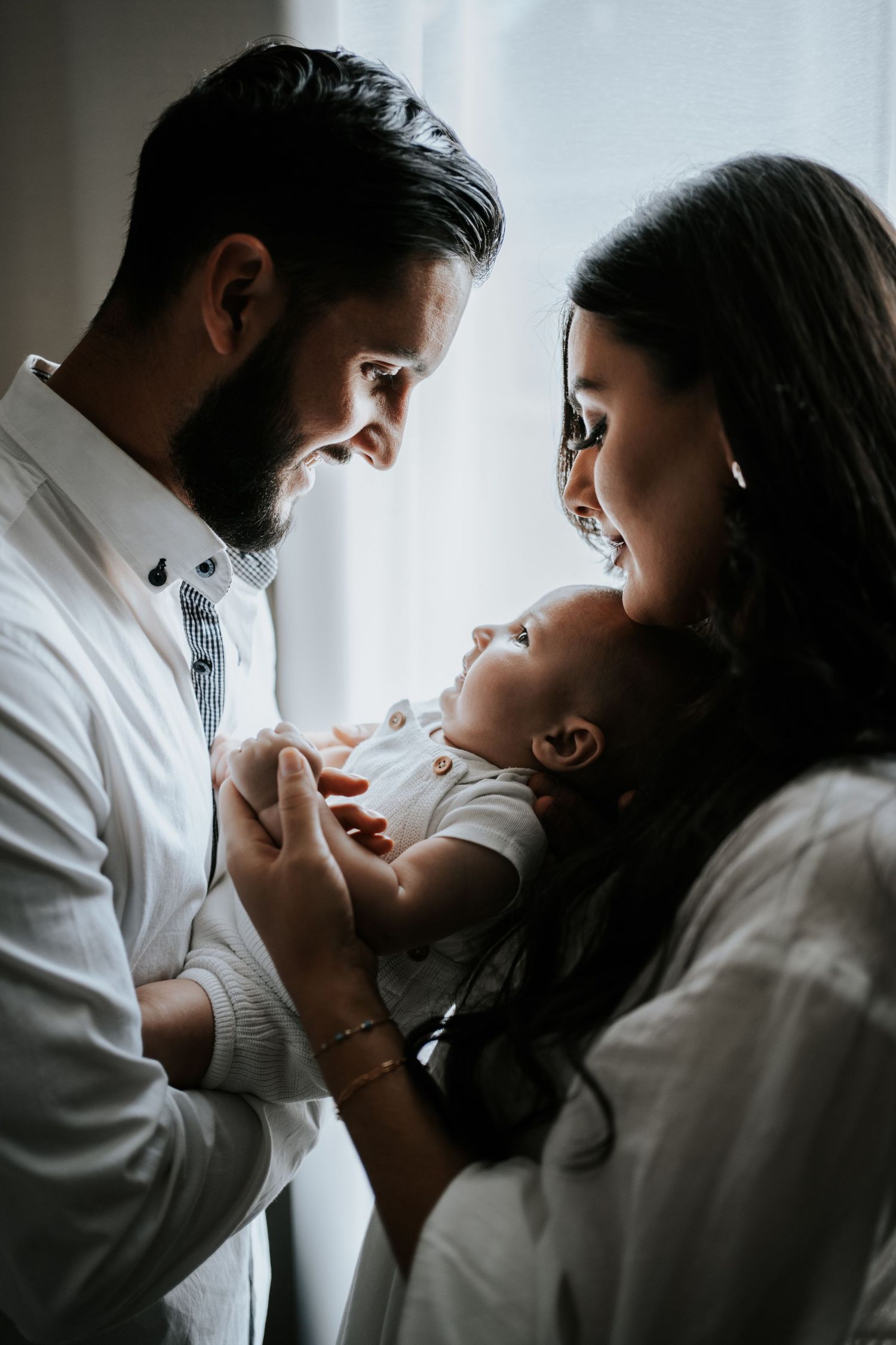 Bearded father and mother gaze lovingly at their newborn baby beside a window in soft natural light.
