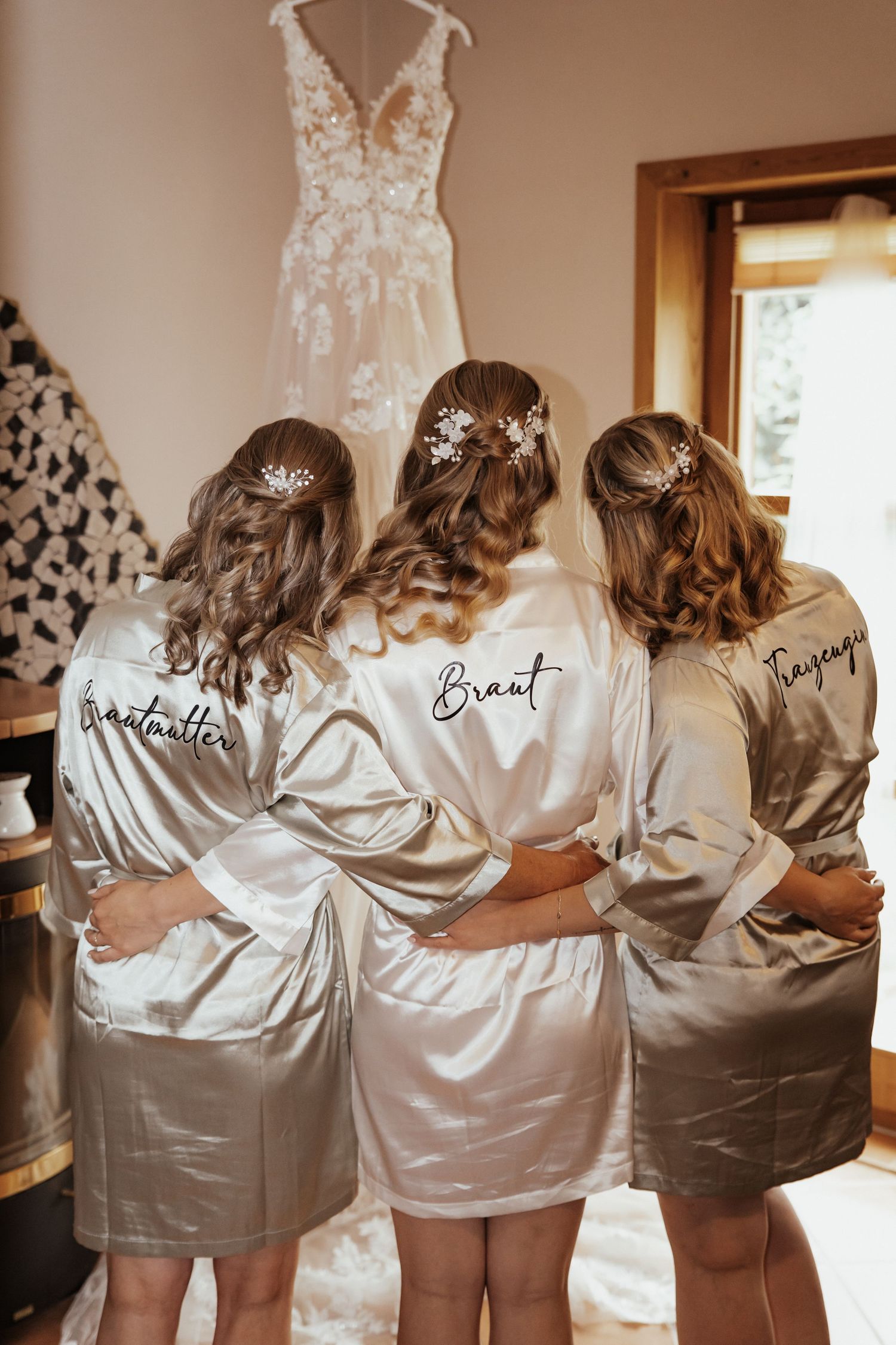 Die Braut und zwei Brautjungfern umarmen sich in Satinmorgenmänteln vor einem aufgehängten Brautkleid aus Spitze.